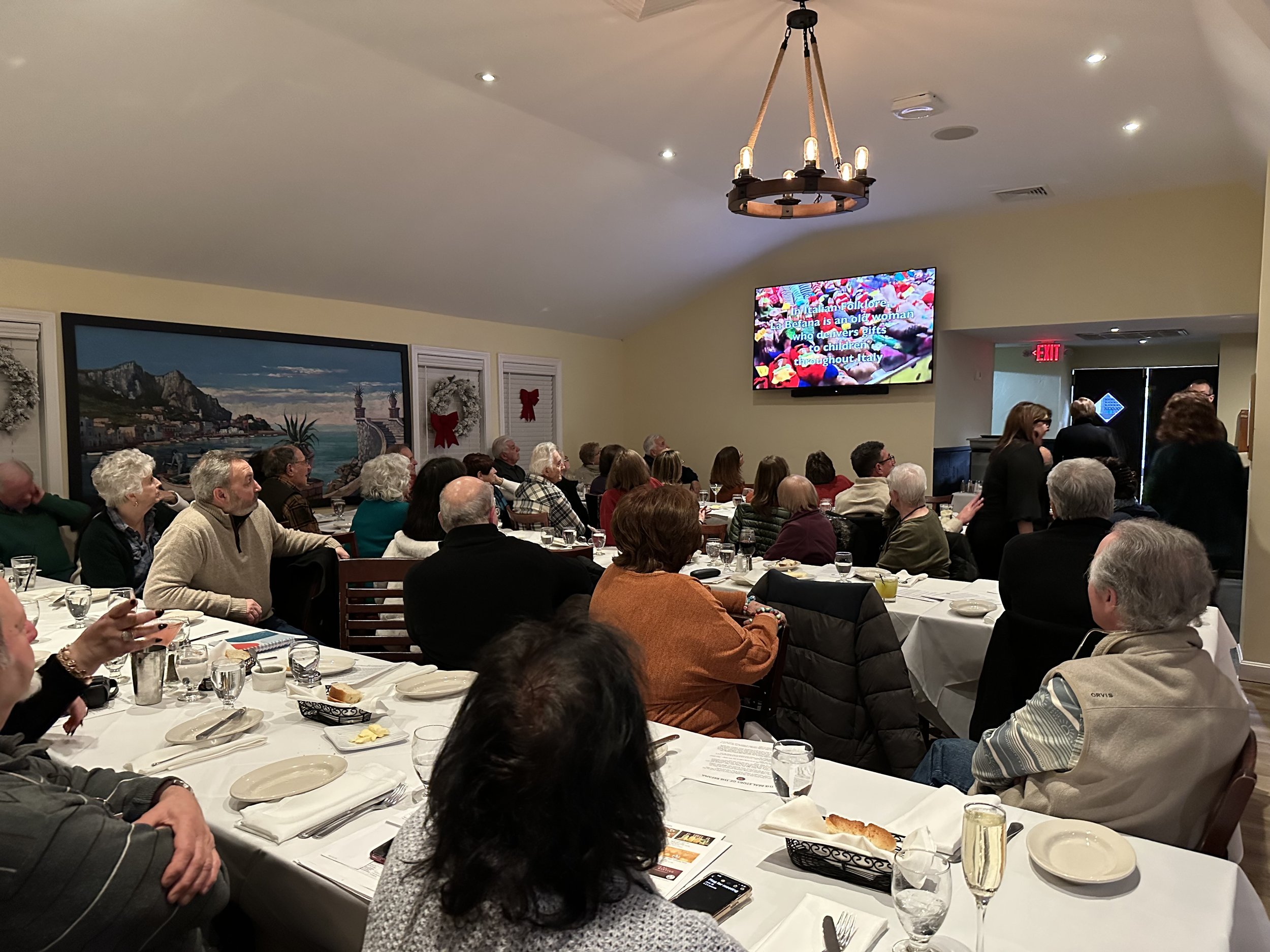 A group of people seated at tables in a restaurant event, watching a presentation on a wall-mounted screen. The room is decorated with paintings and holiday wreaths. Tables are set with white tablecloths, plates, and glasses.
