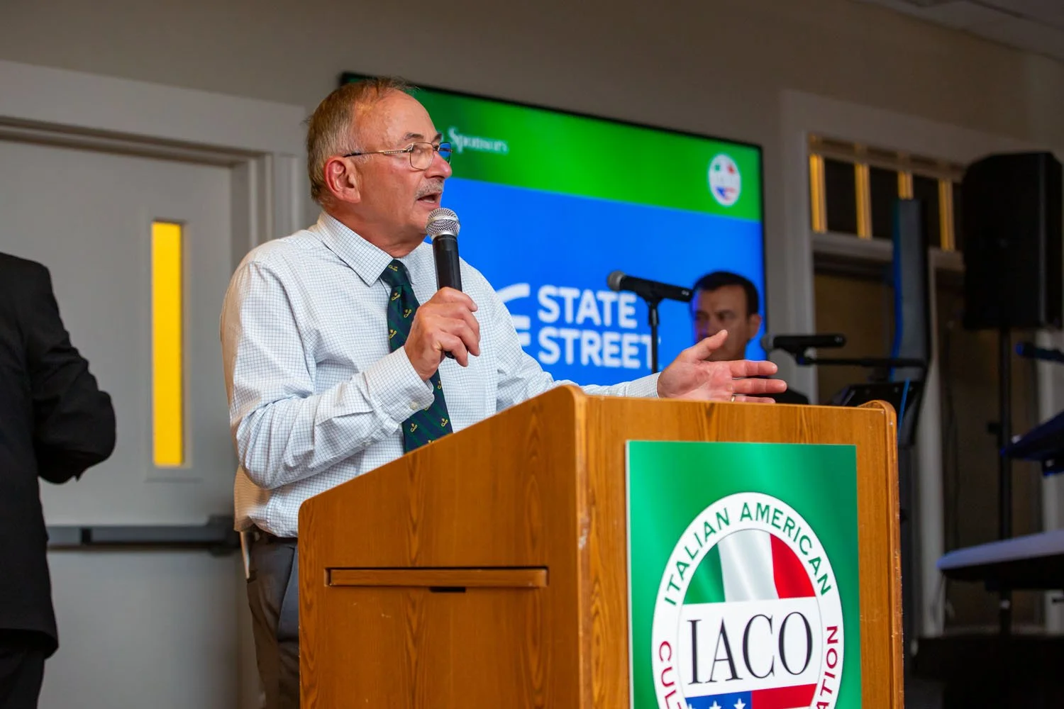 An older man with glasses and a patterned tie speaking into a microphone at a wooden podium with the IACO logo, in front of a large screen with the words 'State Street' and the Italian American Civic Organization emblem.