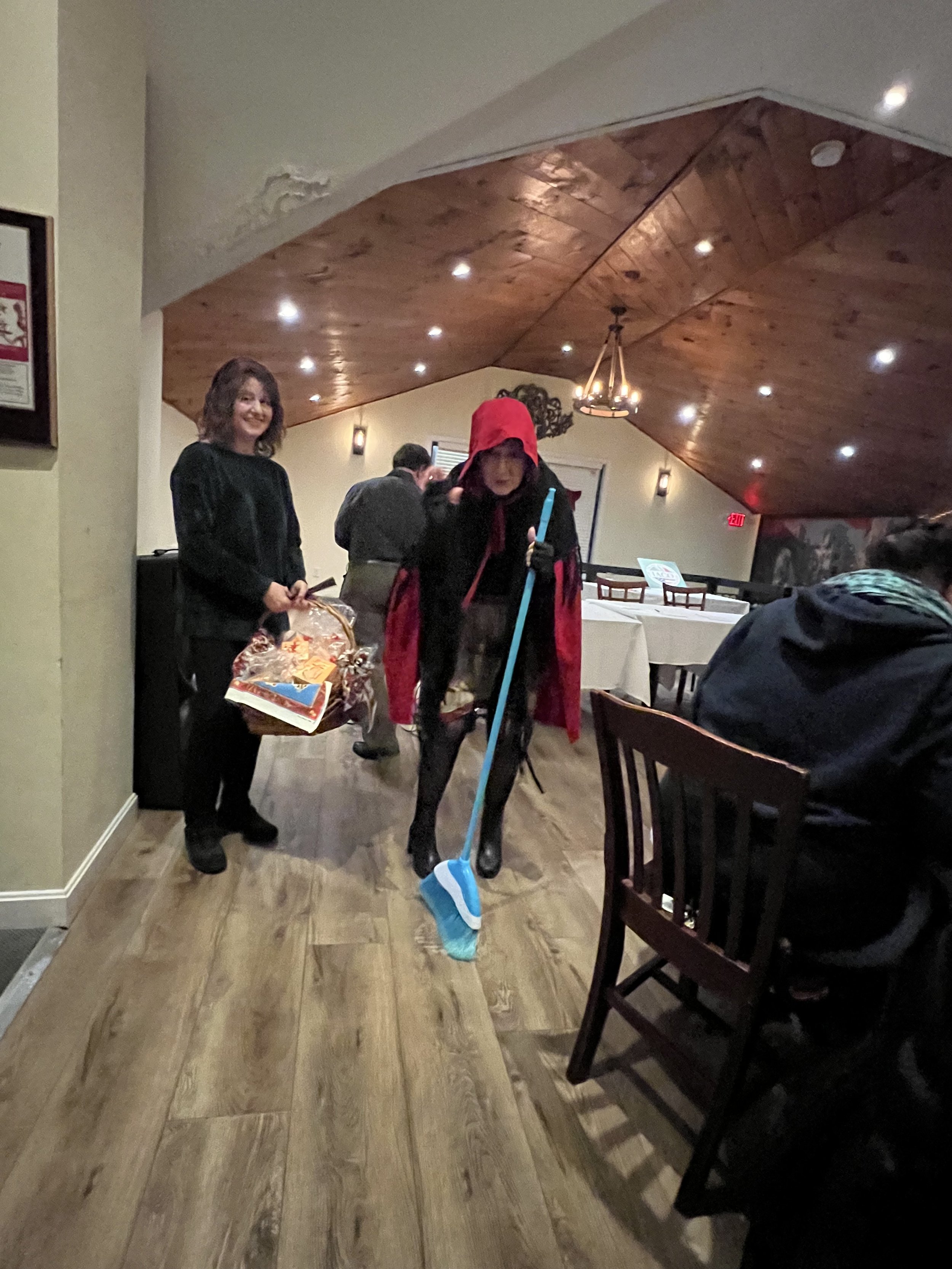 A person wearing a red hooded cape walking indoors, holding a blue broom, with a woman smiling nearby holding a decorative bag. The setting appears to be a room with wooden flooring and ceiling, and dining tables in the background.