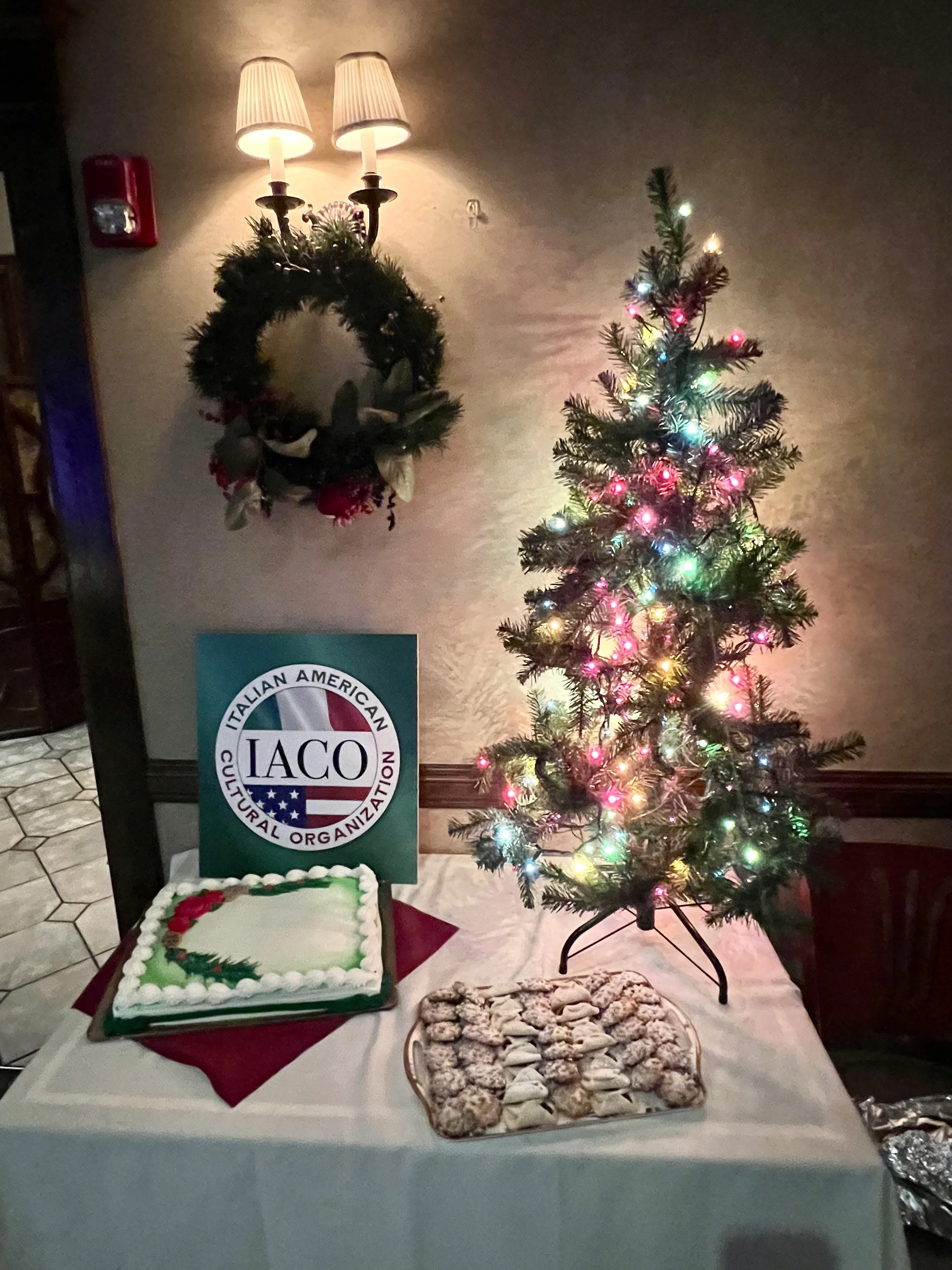 A small decorated Christmas tree with colorful lights on a table, with a holiday-themed cake, a tray of cookies, and an IACO sign. A holiday wreath hangs on the wall above the table, and a wall-mounted lamp is lit.