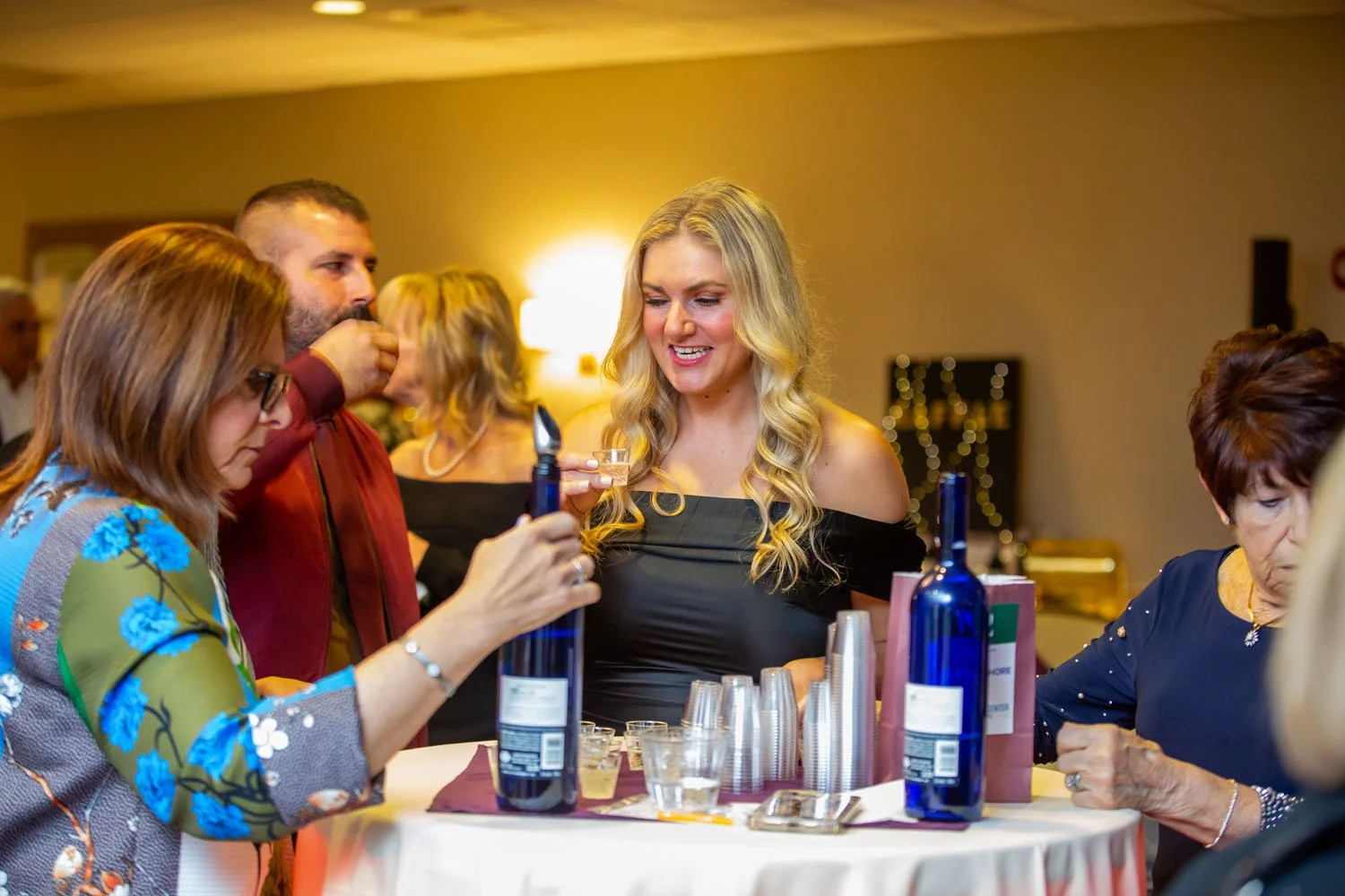 People gathered at a social event, standing around a table with drinks and bottles, engaging in conversation.