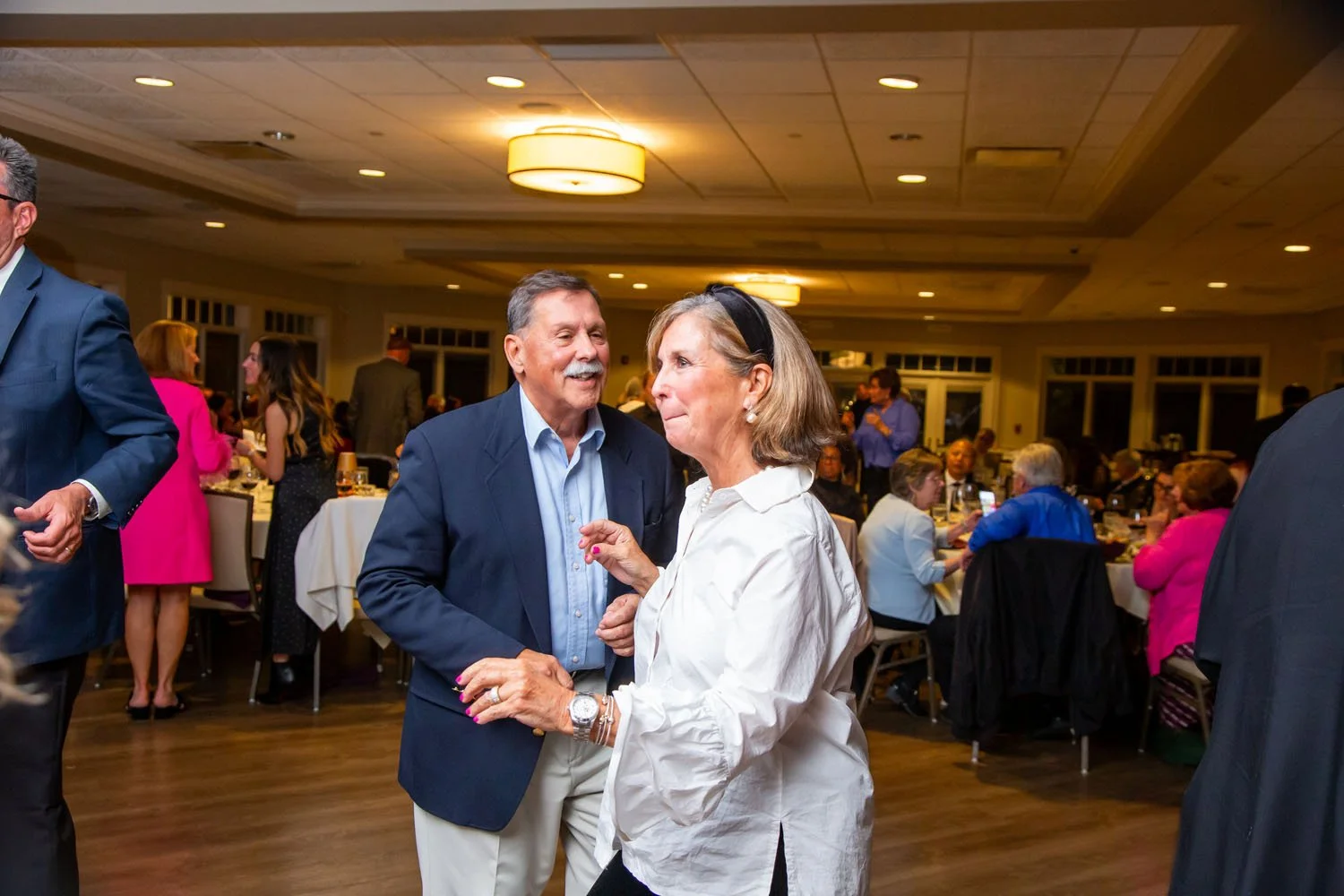 An elderly man and woman dancing and talking at a social event in a ballroom. The woman is wearing a white shirt and pearl earrings, and the man is in a blue blazer. There are other people dining and socializing in the background.