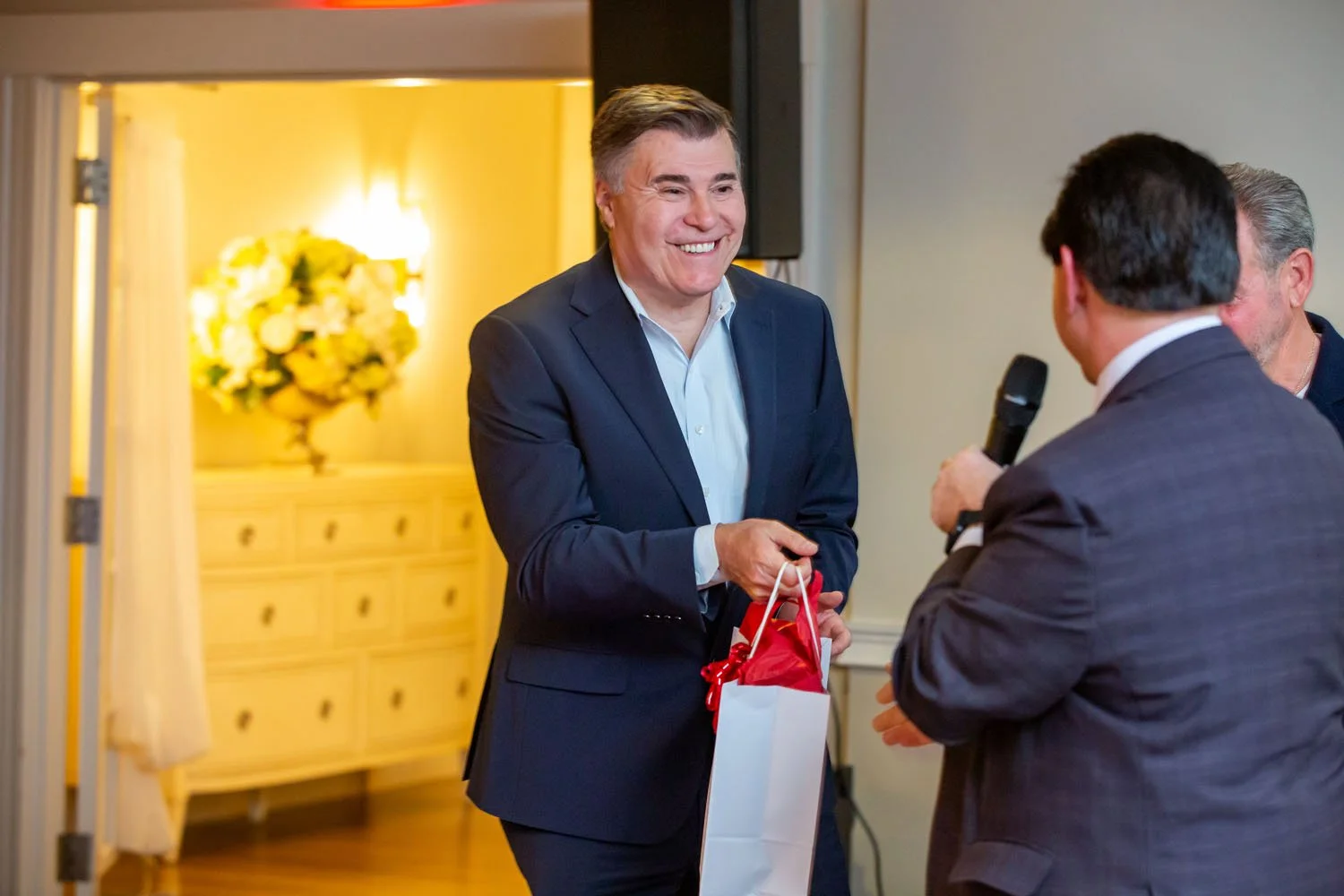 Man in a dark suit smiling and exchanging a gift bag with another man who is holding a microphone at an indoor event.
