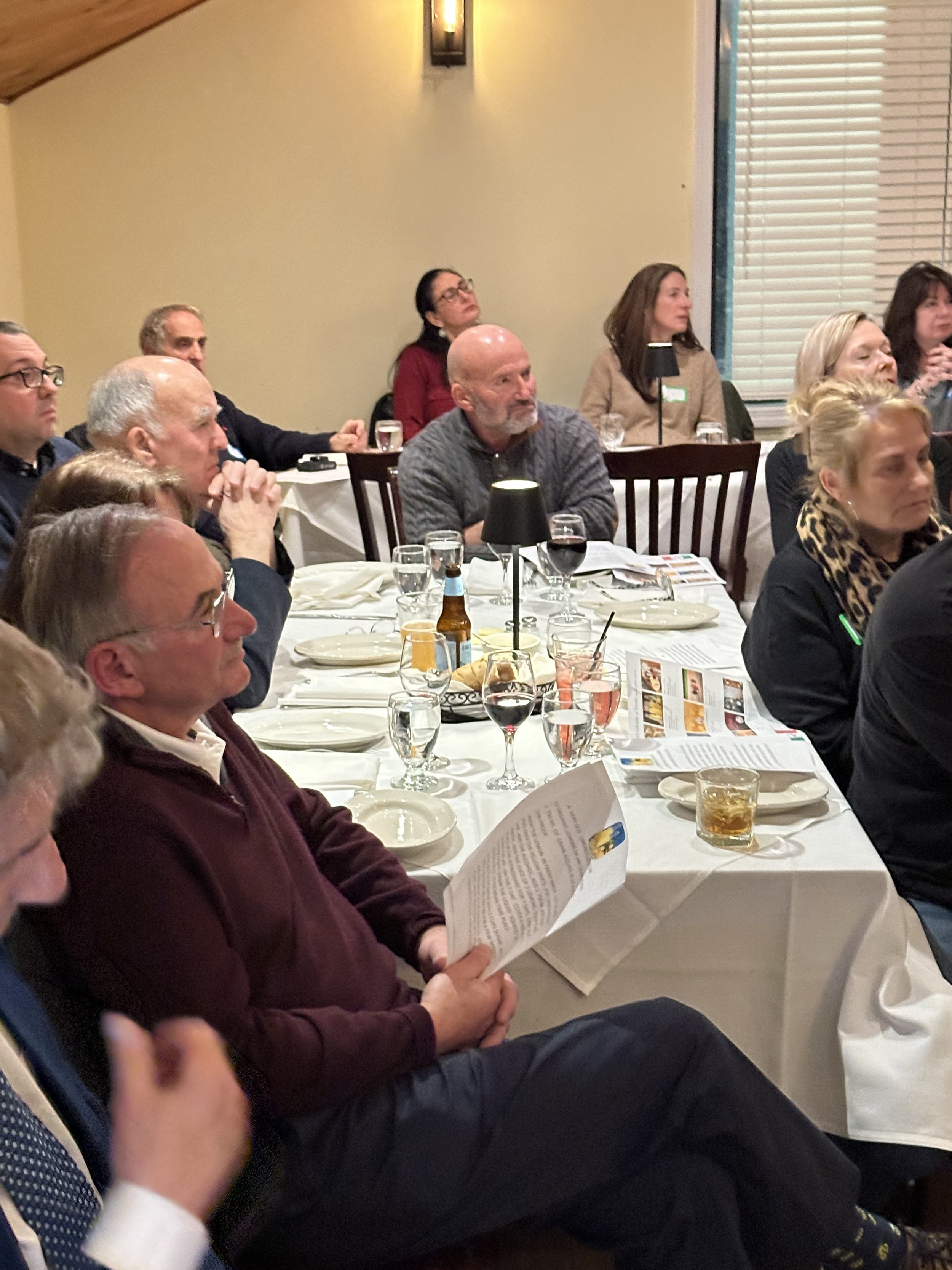 People sitting at tables in a gathering or event, focused on someone speaking or presenting off-camera. Tables are set with drinks and papers.