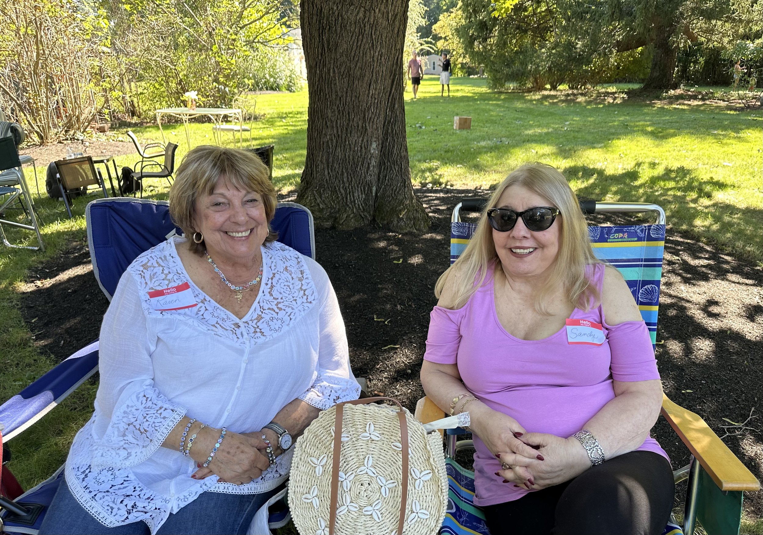 Two women sitting on outdoor chairs under a tree, smiling. They are wearing name tags and casual clothing. The setting appears to be a park with grass, trees, and additional chairs in the background.