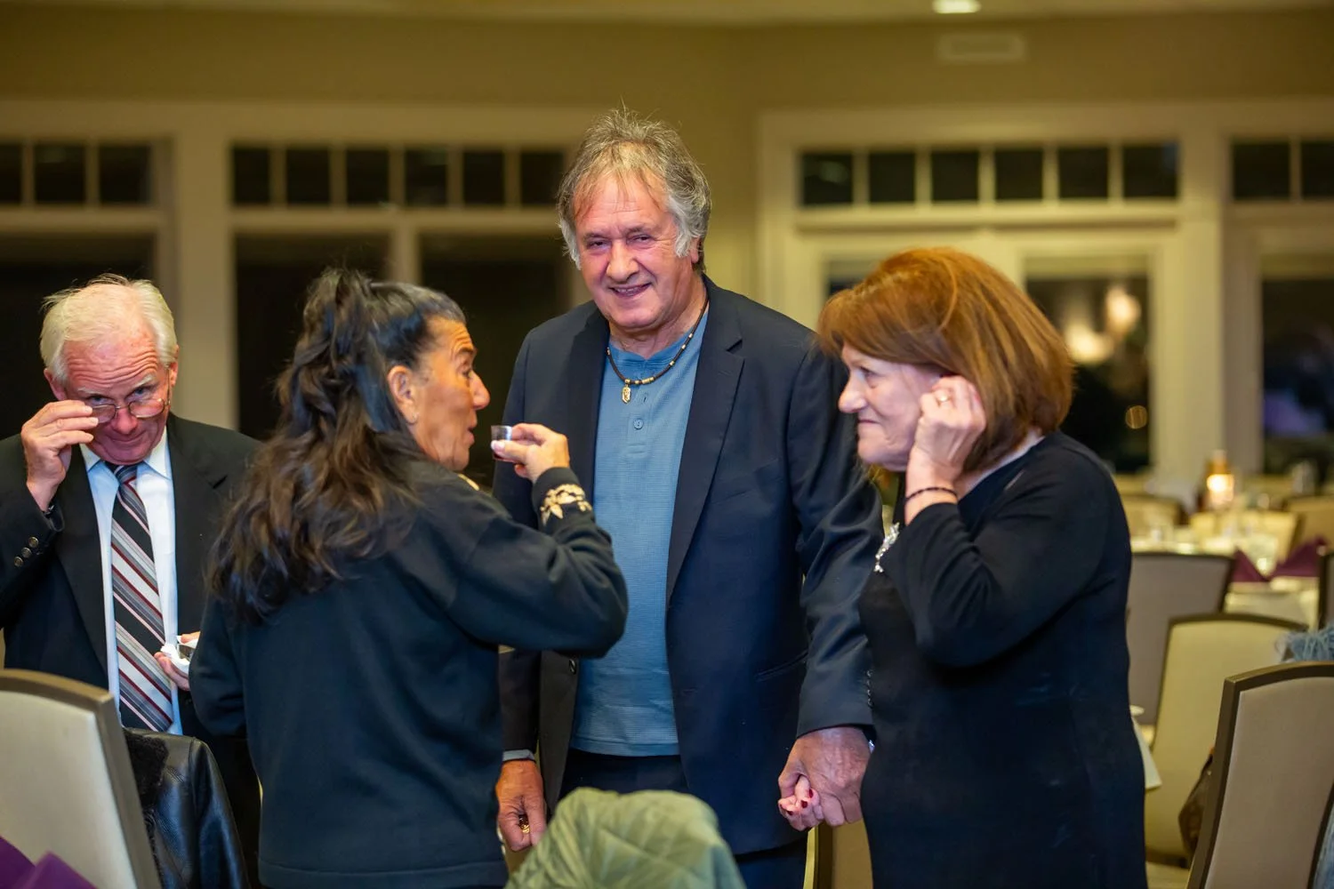 Four people at a social gathering in a restaurant or banquet hall. One woman is handing a small glass to a tall man in the center, all four are engaged in conversation and smiling. The background shows large windows and tables.