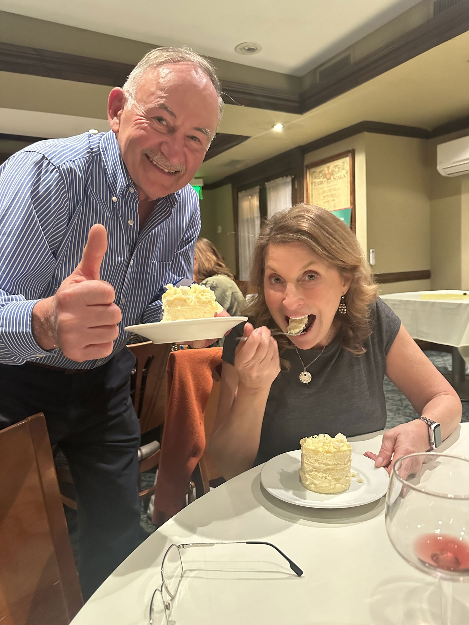 A man and a woman are enjoying dessert at a restaurant. The man is giving a thumbs up and holding a plate with a cake slice, while the woman is eating a piece of cake. There are two plates with cake slices on the table, and a glass of red wine is vis