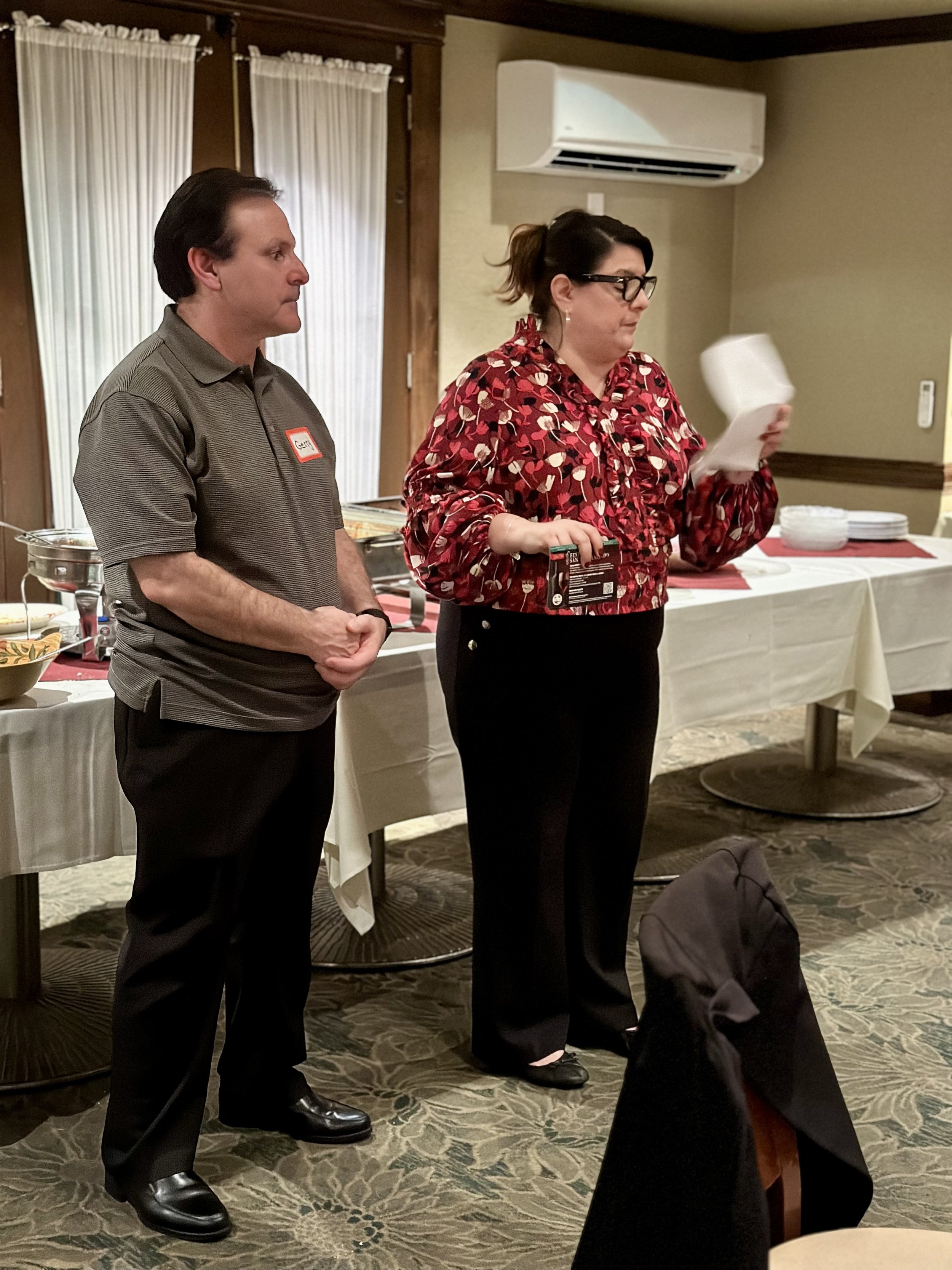 Two people standing in a room with buffet tables and air conditioning unit in the background; one person holding papers.