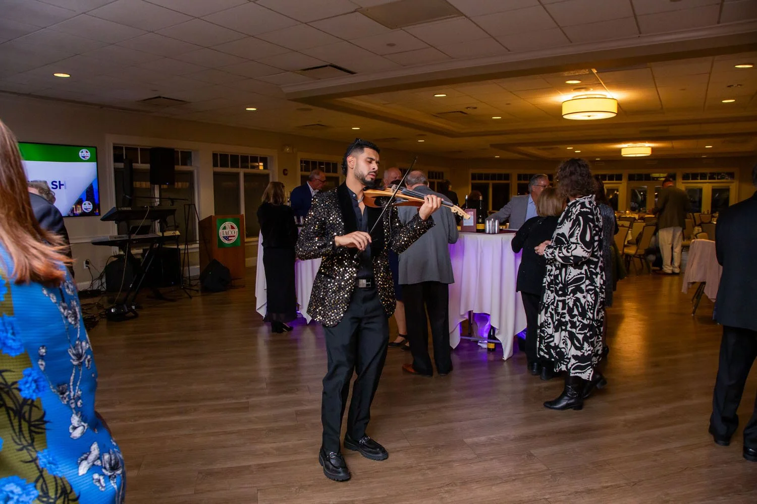 A man in a black sequin jacket playing the violin at a social gathering indoors with other guests standing and talking around him.