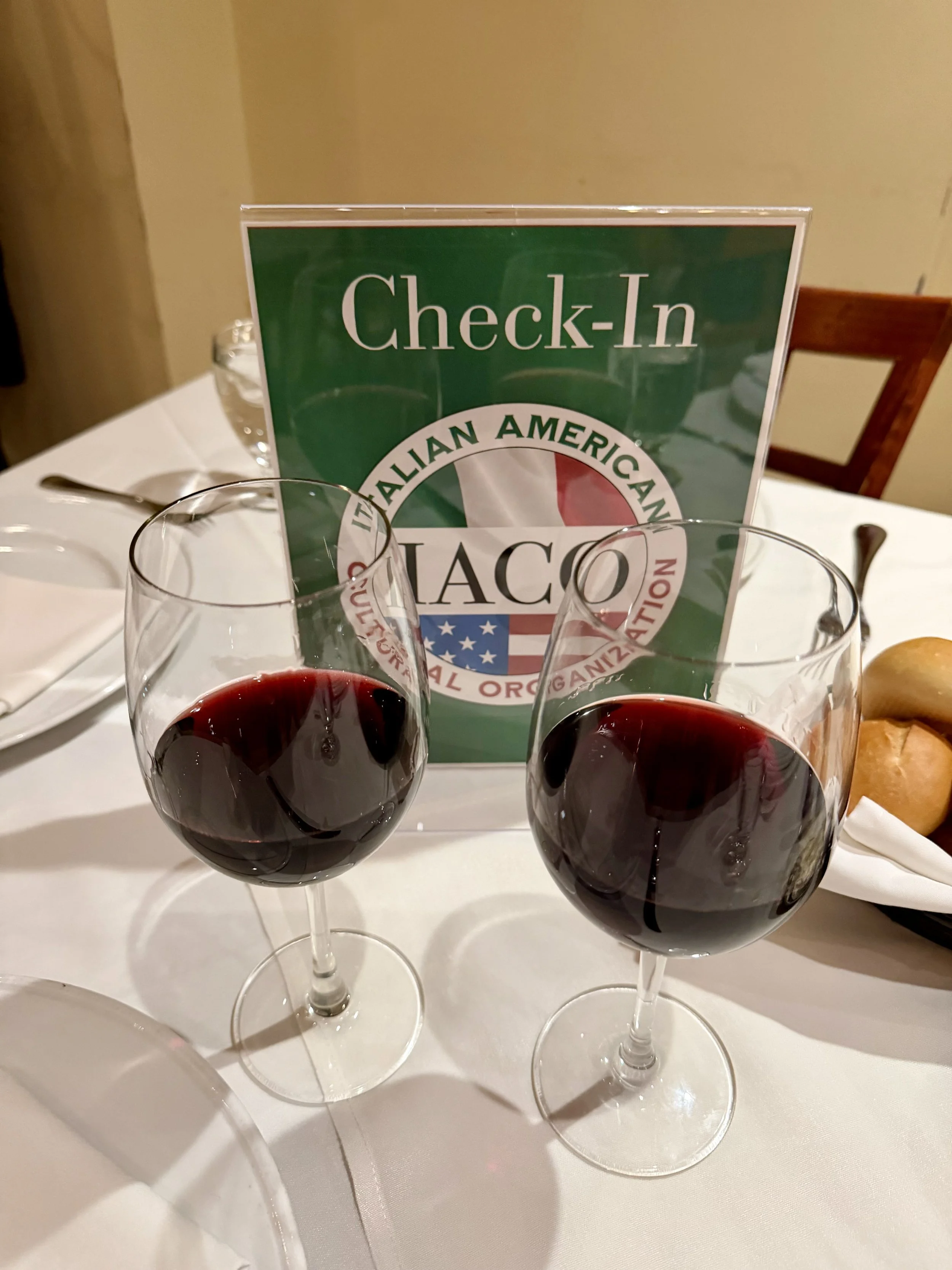 Two glasses of red wine on a white tablecloth with a check-in sign for Italian American TACCO restaurant in the background.