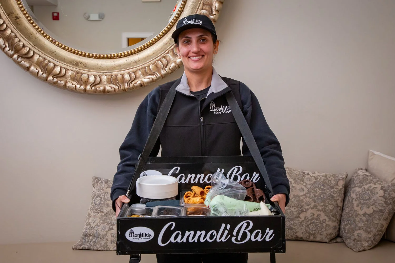 A smiling woman wearing a black Montilio's Baking Company cap and jacket holding a Cannoli Bar tray with assorted items inside. She stands in front of a beige wall with a large, ornate gold-framed mirror and decorative pillows on a sofa.