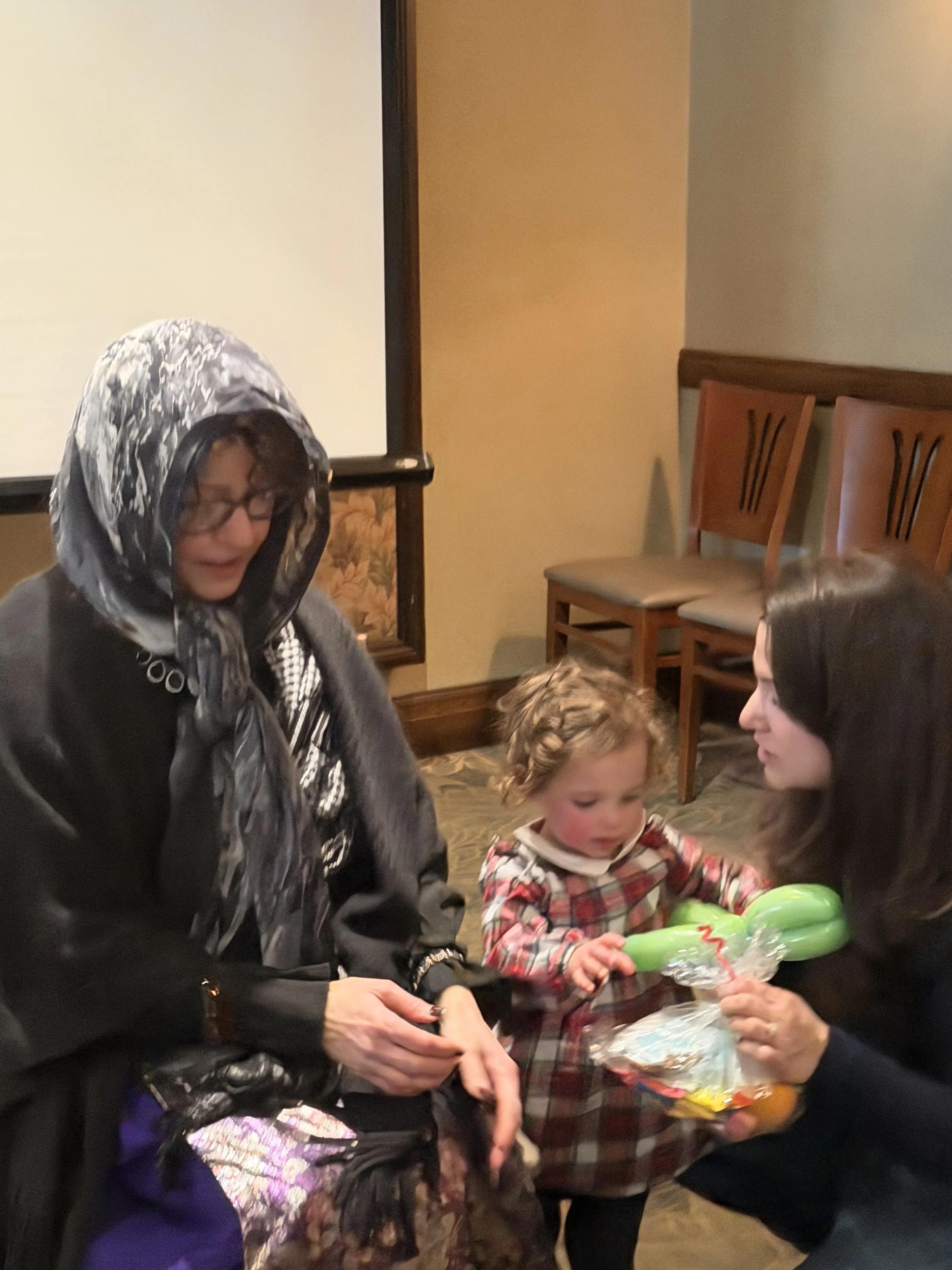 Three people, a woman with glasses and a scarf, a young girl with curly hair in a checkered dress, and another woman with dark hair, are sitting around a table. The young girl is holding a green balloon animal, and the woman on the right is offering 