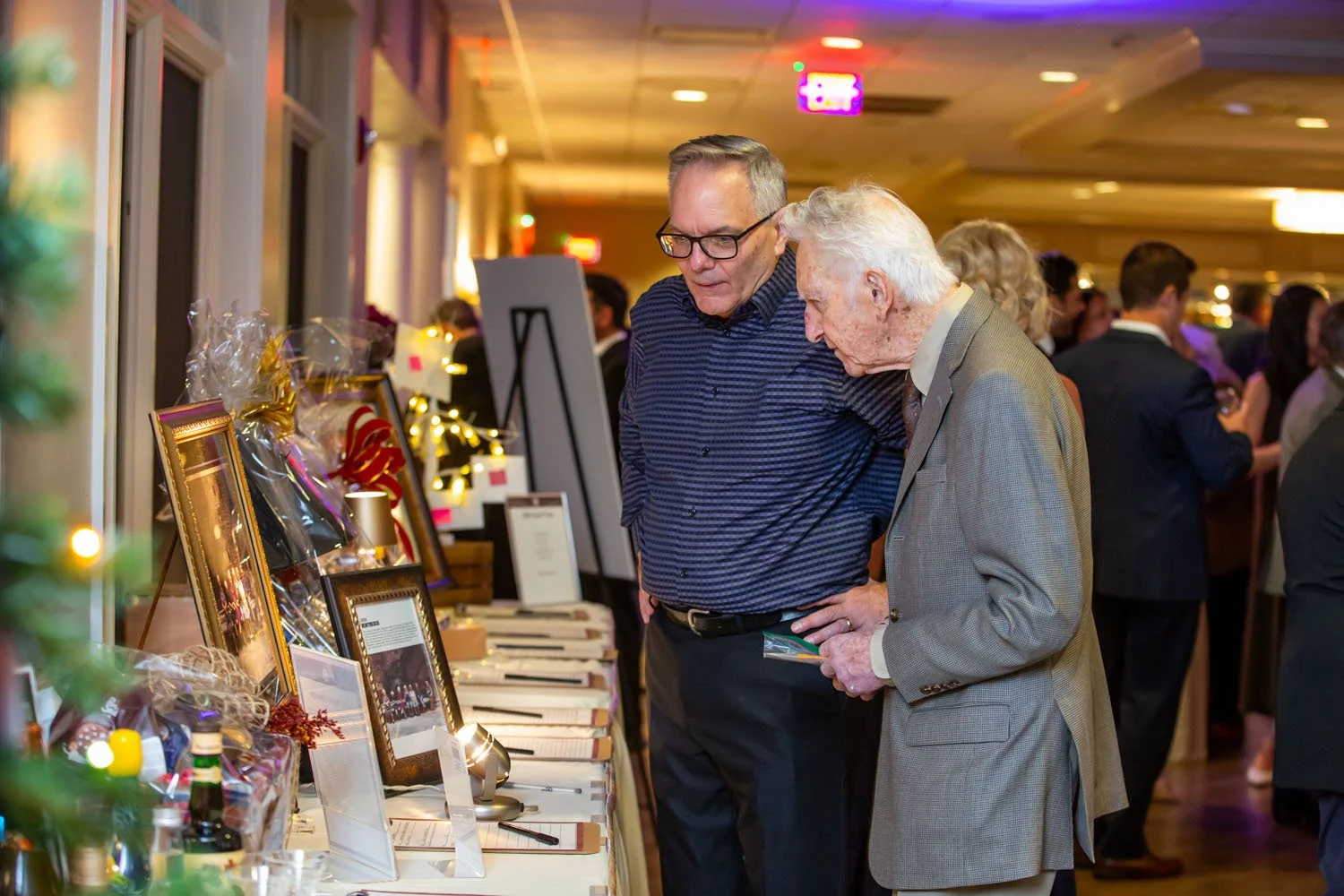 Two men, one elderly and one middle-aged, look at items on a display table at an indoor event, surrounded by other guests.
