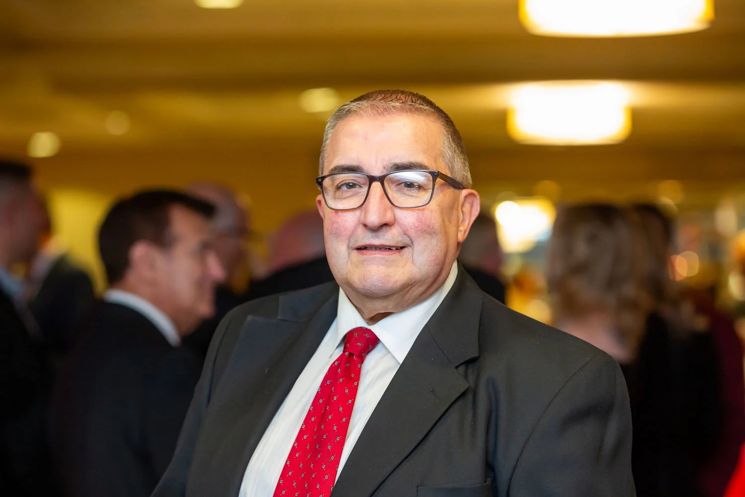 A middle-aged man with glasses, wearing a dark suit, white shirt, and red tie, standing in a crowded, well-lit event space.
