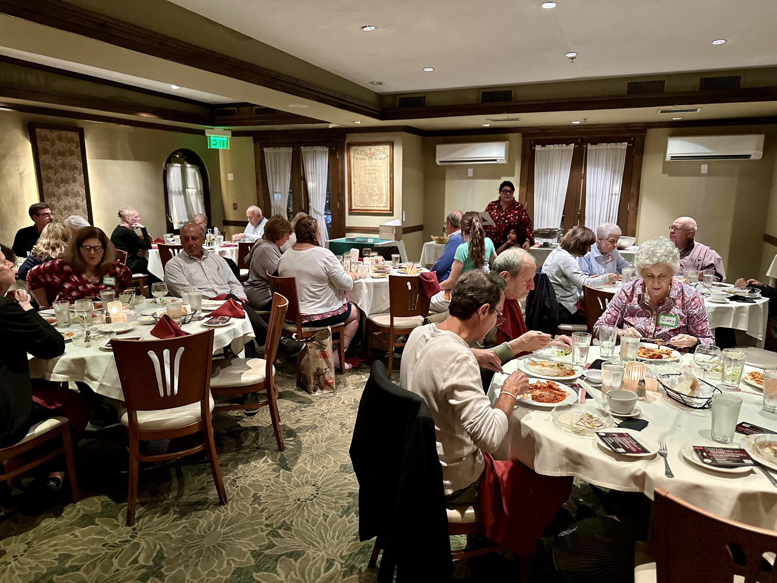 People dining in a restaurant with round tables set with plates and glasses, engaged in conversation and eating.