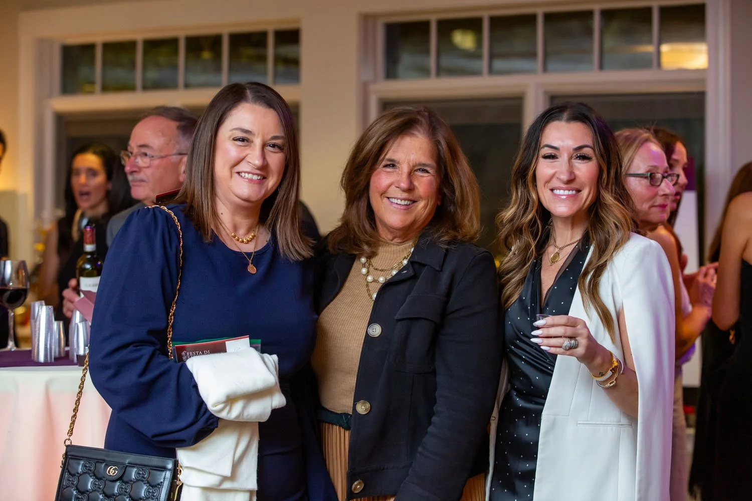 Three women smiling at a social event, standing close together, with other guests and a table with drinks in the background.