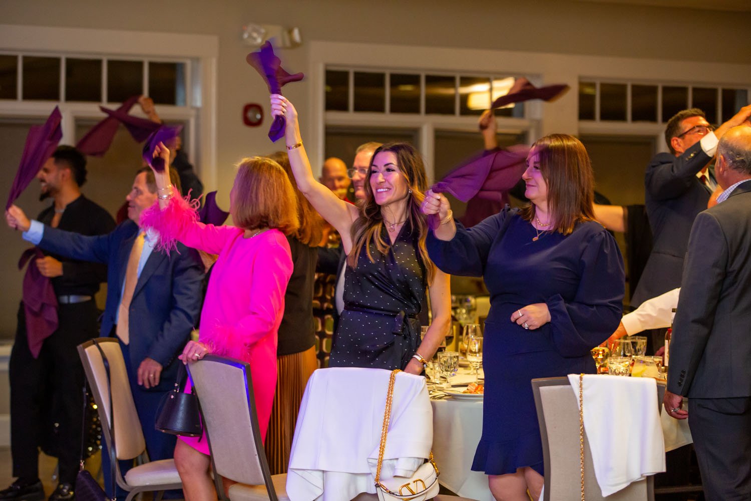 People celebrating at a formal event, raising napkins, standing around a dinner table with glasses and cutlery, in a well-lit room.
