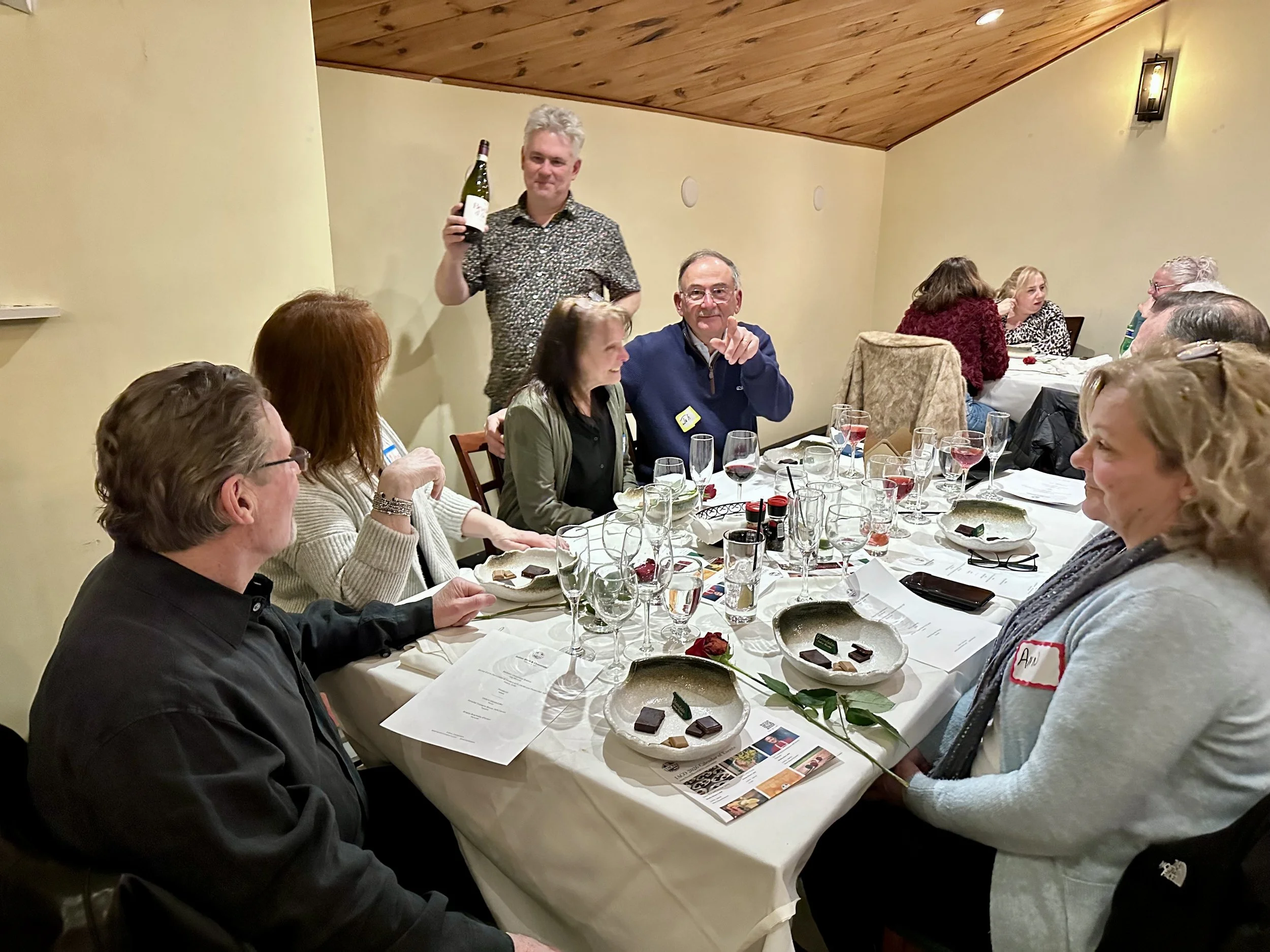 People sitting around a dinner table at a social gathering, with a man standing holding a wine bottle and smiling, others engaged in conversation, glasses of wine and plates of food on the table, some people wearing name tags, in a room with wooden c