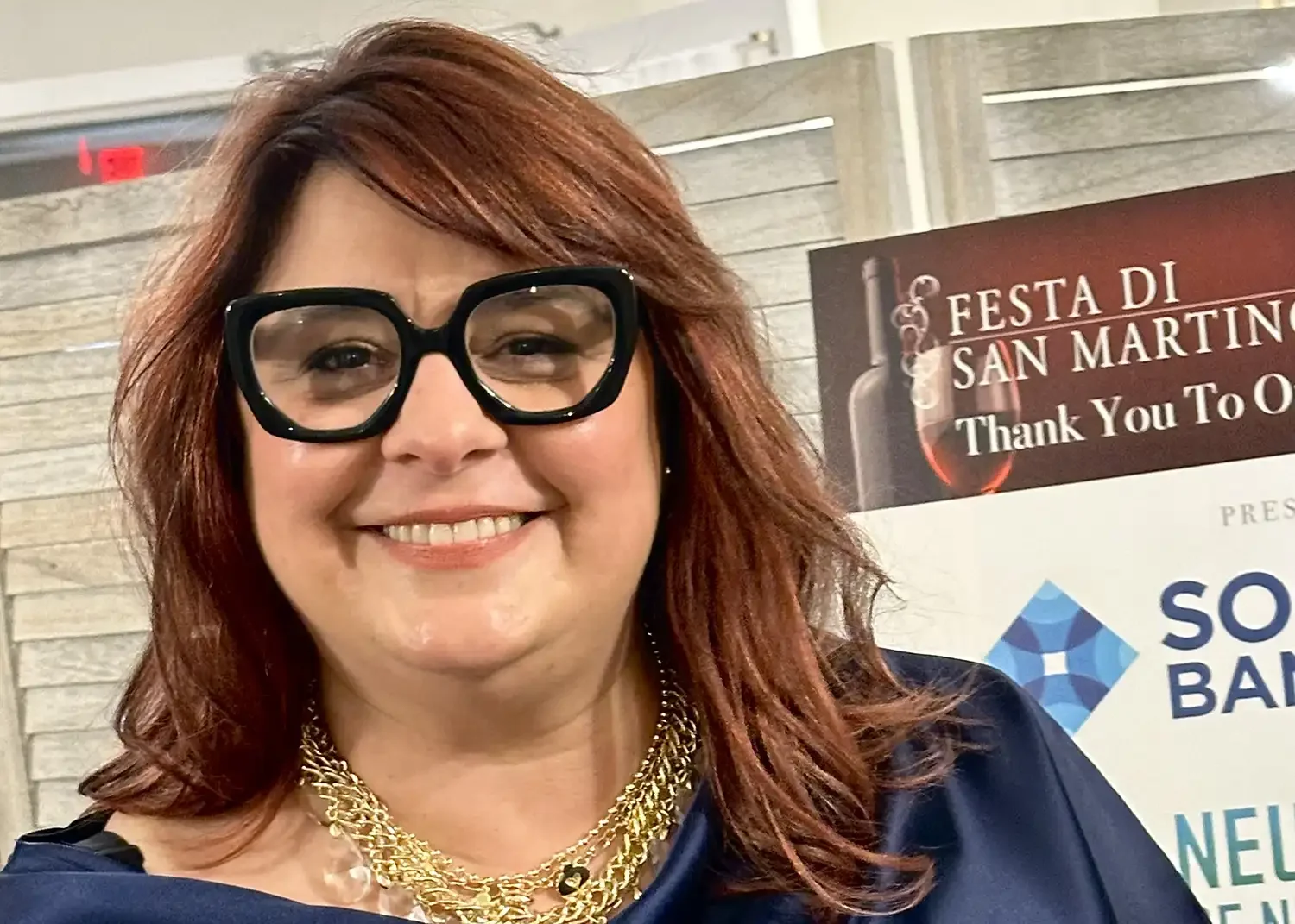 A woman with red hair wearing large black glasses, a blue satin top, and multiple gold necklaces, standing at an event with a sign in the background that says "Festa di San Martino" and "Thank You To of Sponsors".