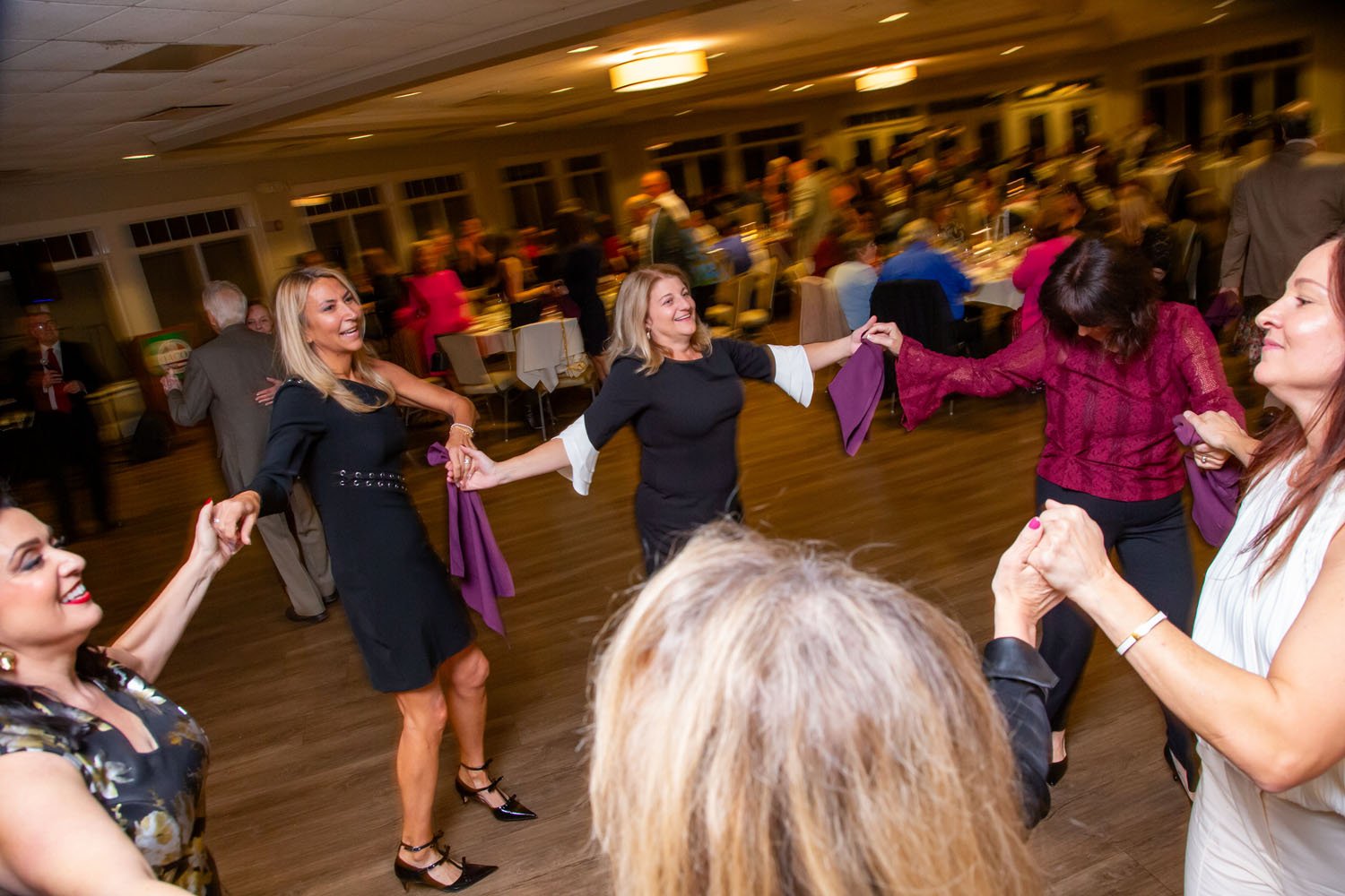 People dancing and holding hands in a circle at a social event or celebration in a banquet hall.