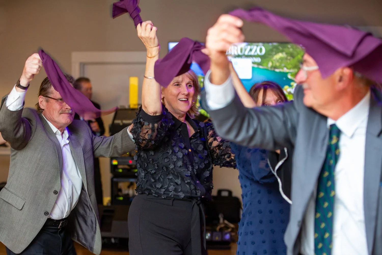 People are dancing together at a celebration, holding purple cloths in a room with a television and audio equipment in the background.