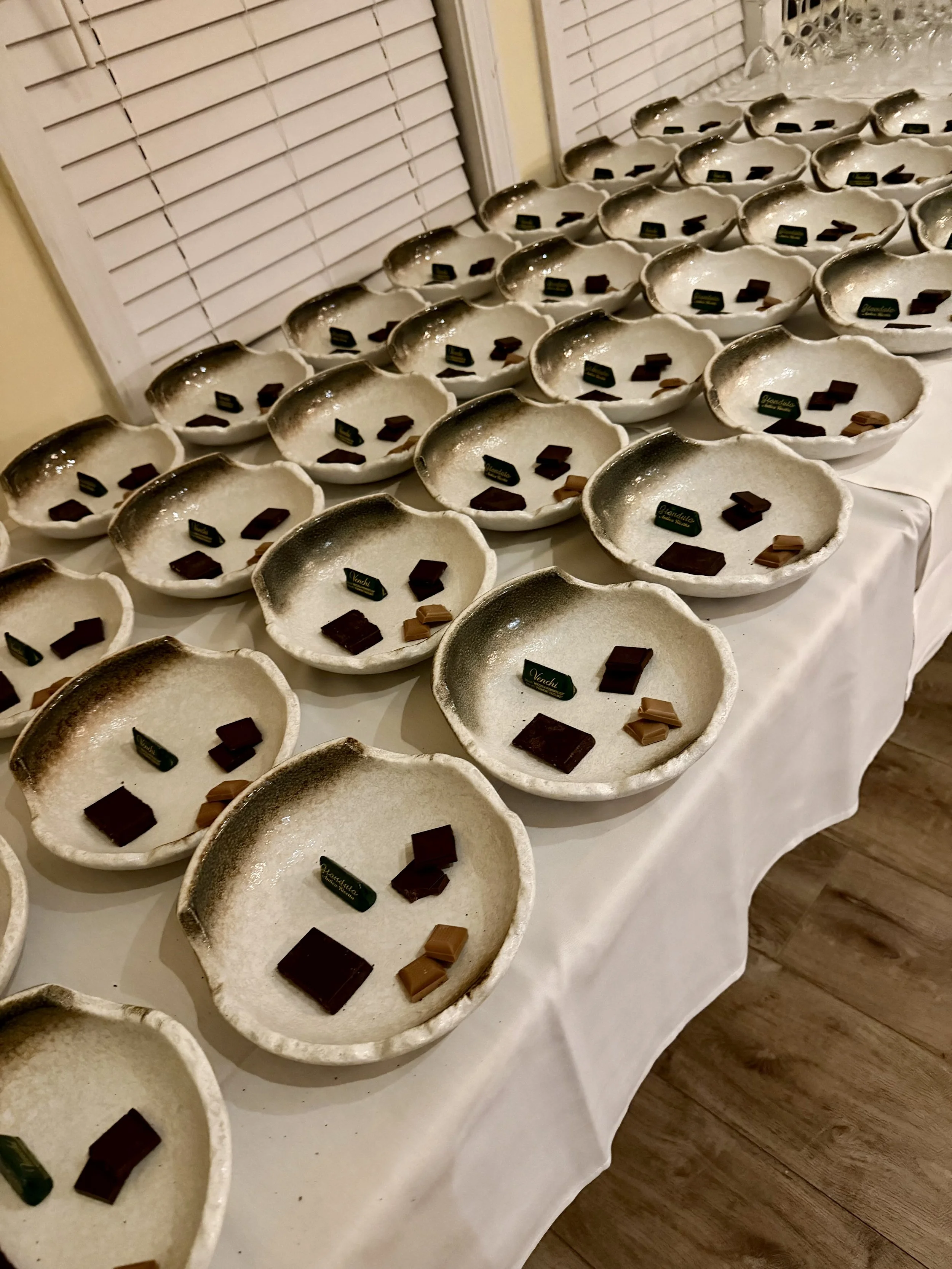 Multiple white ceramic bowls displaying various pieces of chocolate, arranged on a long table covered with a white tablecloth, with closed window blinds in the background.