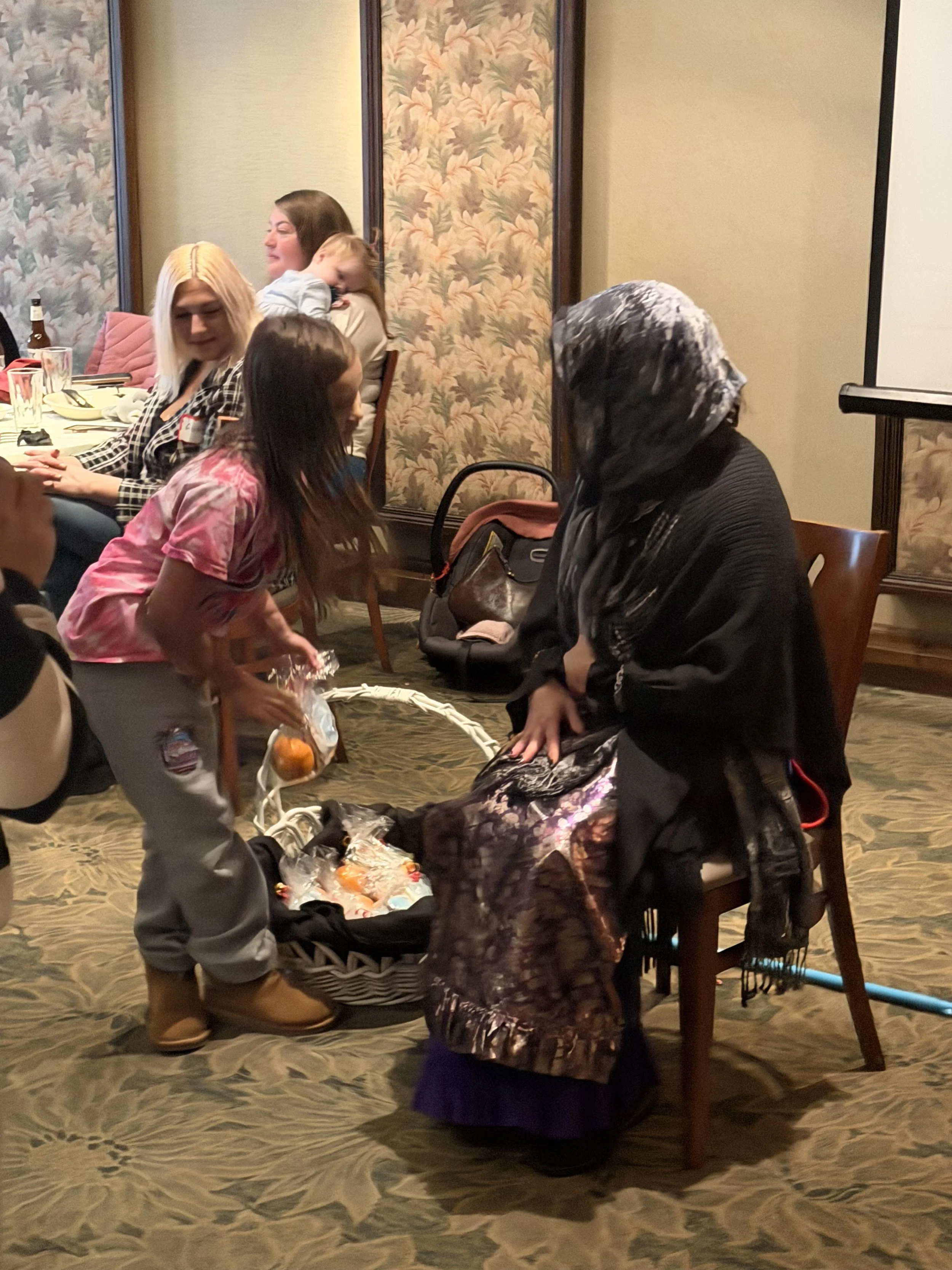 A young girl offering a gift basket to an older woman wearing a headscarf and black clothing, with other women and children sitting at a table in the background.