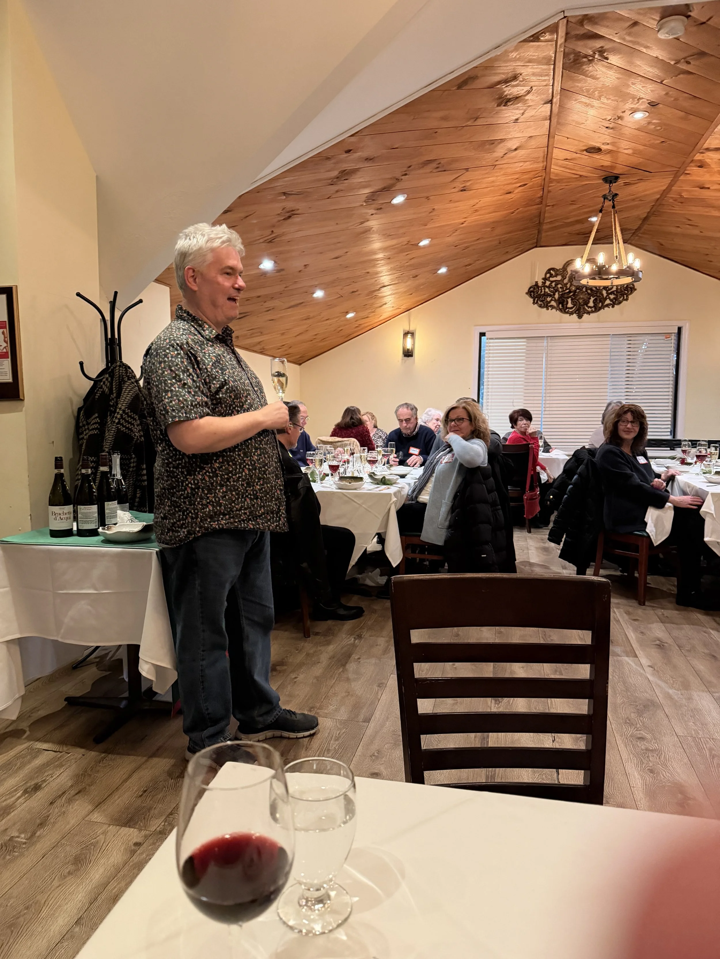 A man standing at the end of a dining table holding a glass of champagne, surrounded by guests seated at tables in a warmly lit restaurant with wooden ceiling and walls.