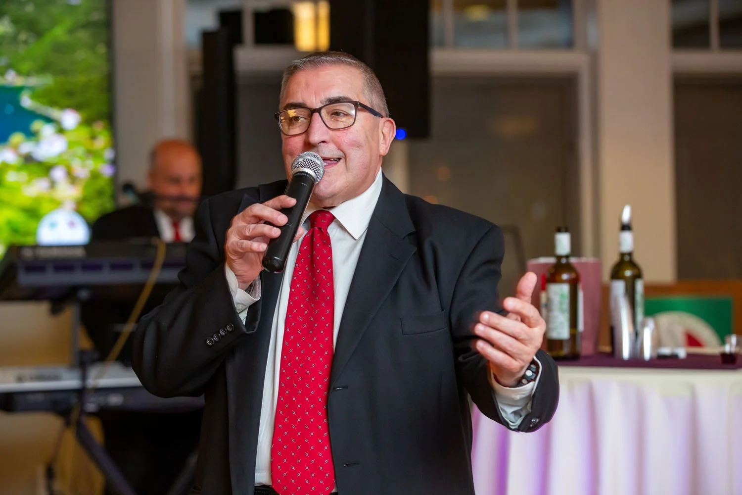 A man in a suit and red tie speaking into a microphone at an indoor event.