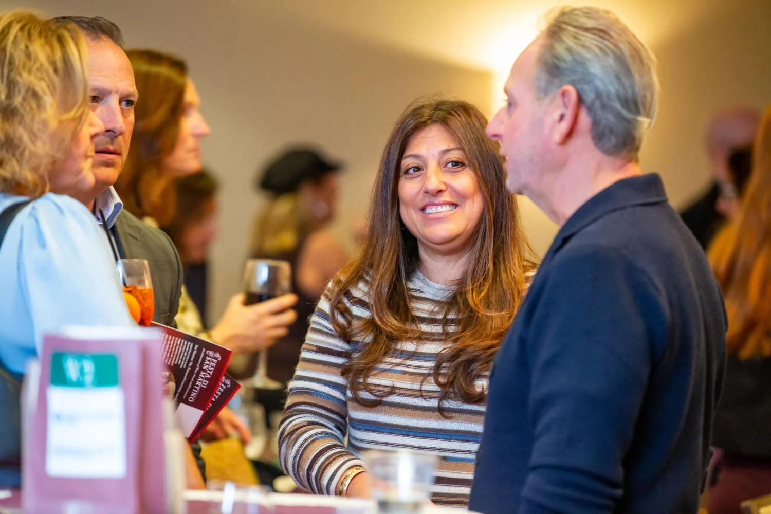 People socializing at an indoor networking event, with a woman smiling at a man and others in the background holding wine glasses.