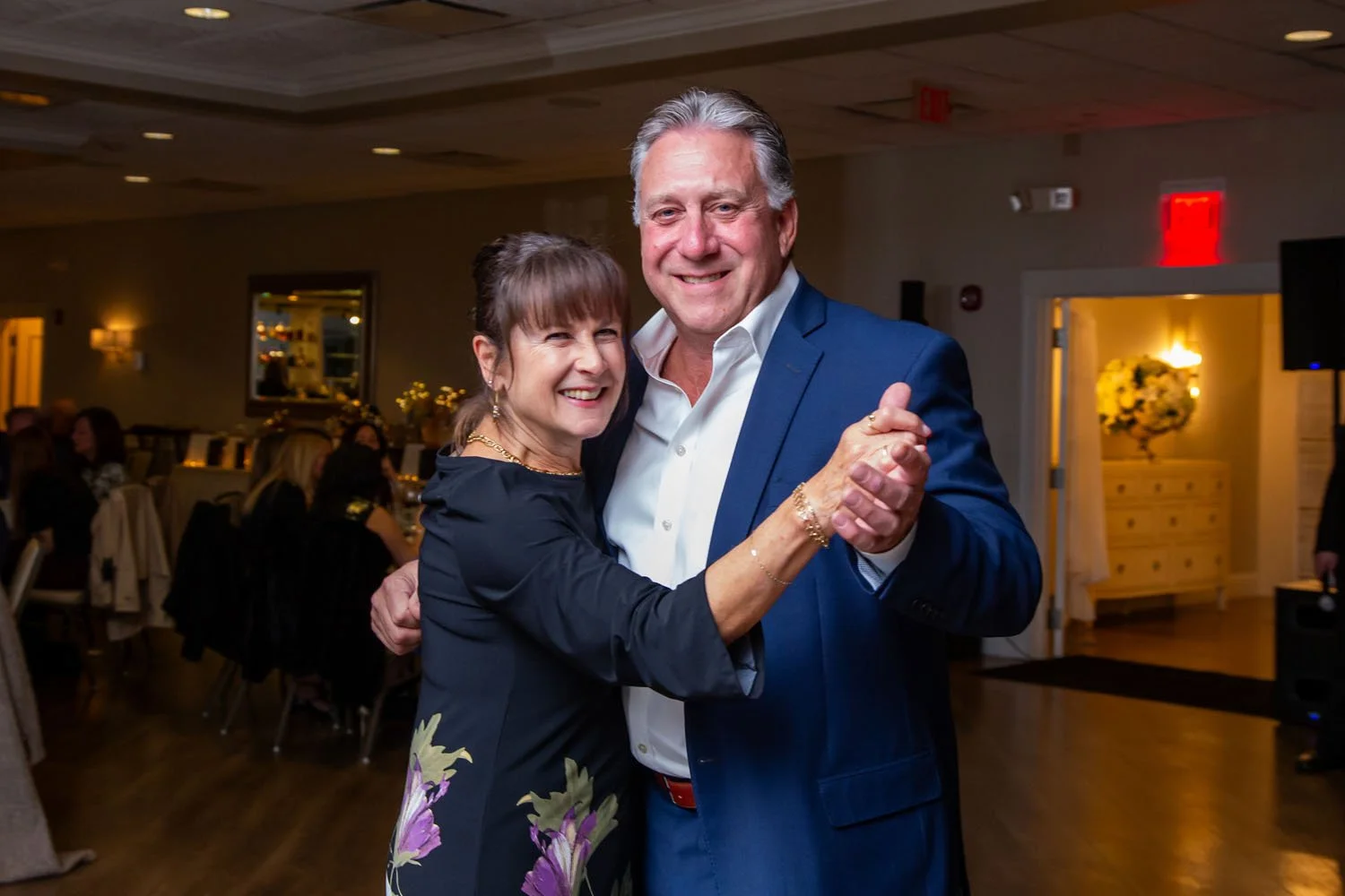 A middle-aged man and woman are dancing together at an indoor event, smiling at the camera. The woman is wearing a dark floral dress, and the man is in a white shirt and blue blazer.