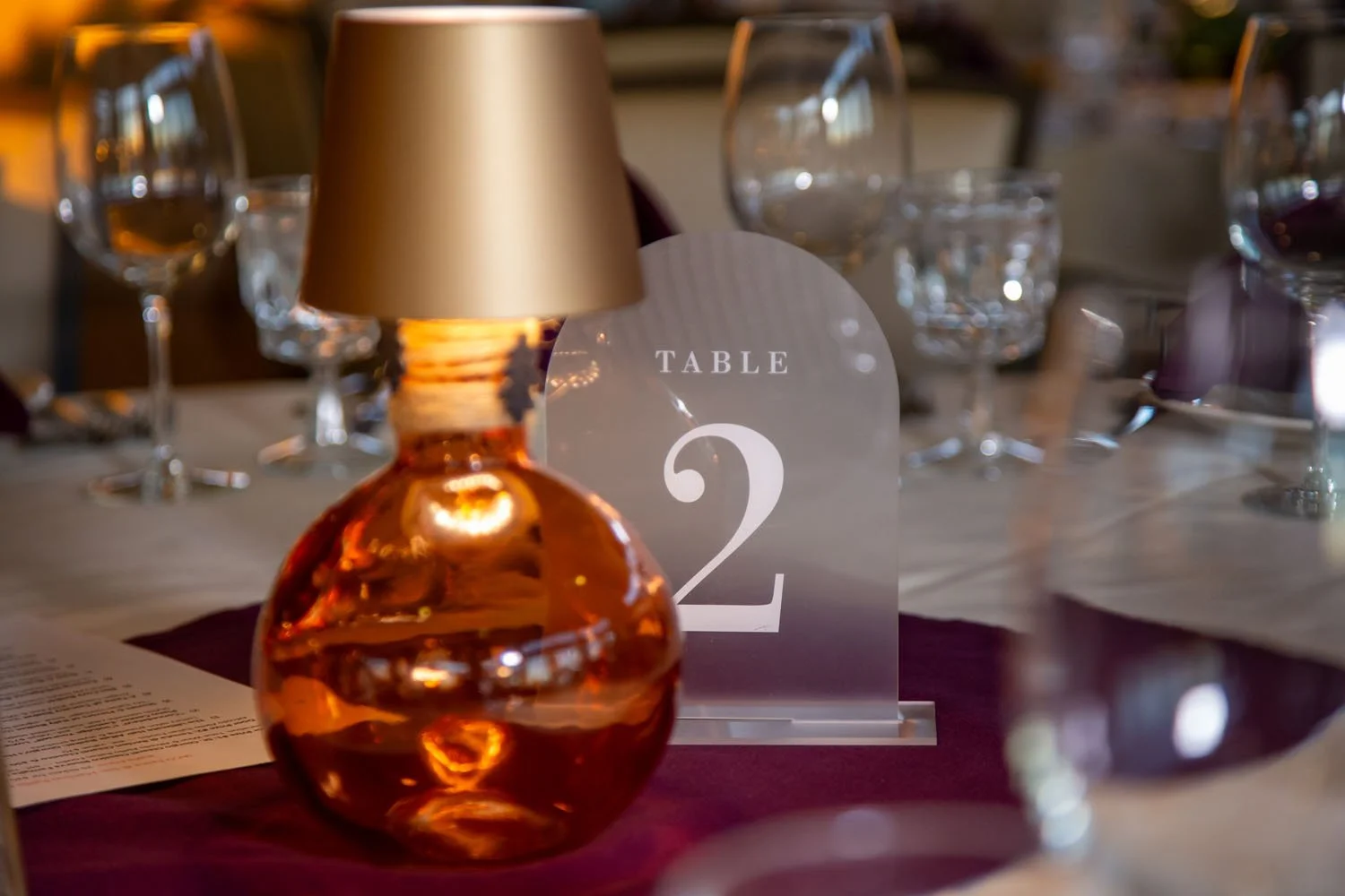 Elegant table setting with wine glasses, a glass table number holder displaying the number 2, and a golden-colored table lamp in a warmly lit dining environment.