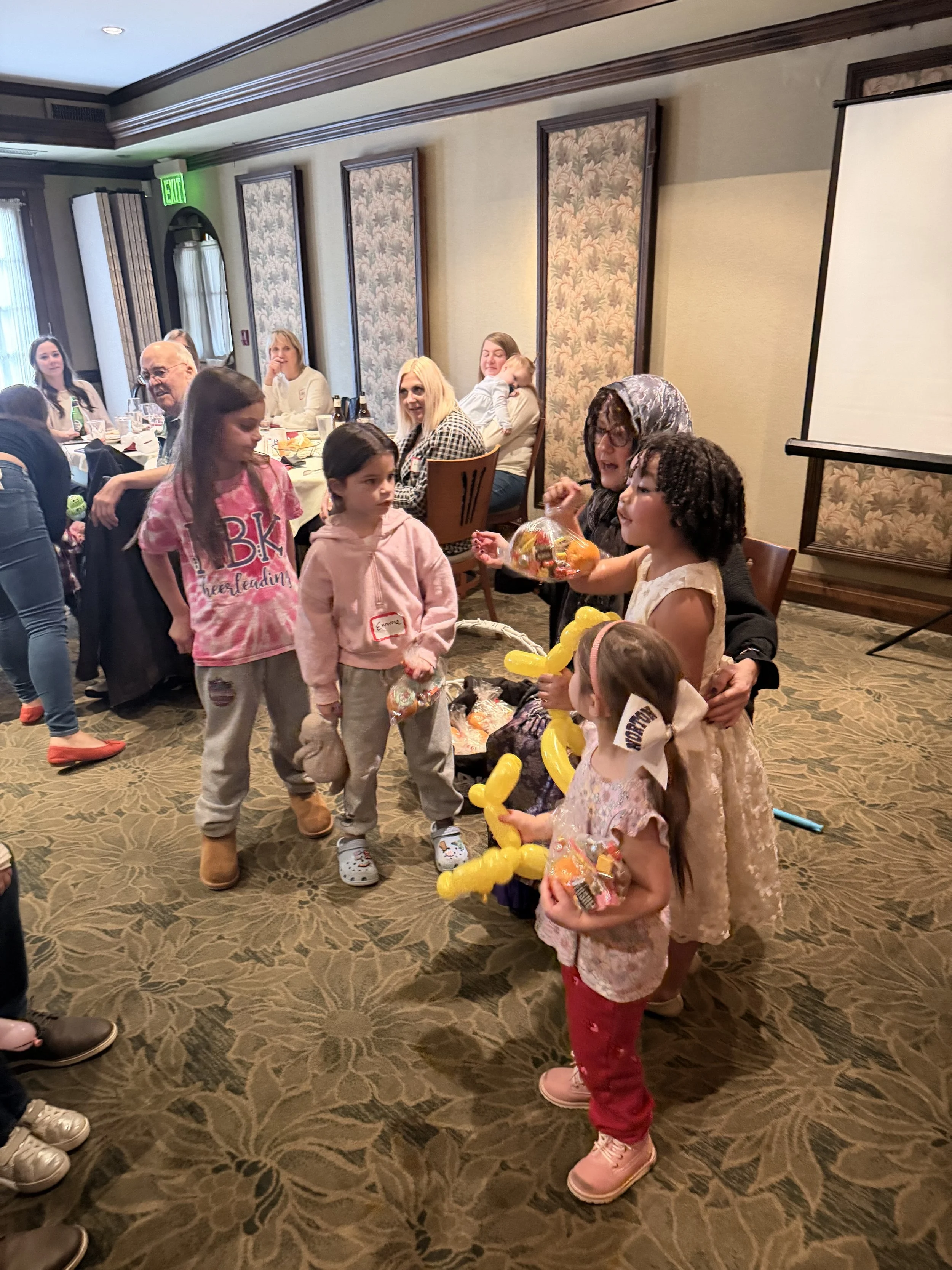 A group of children and an adult woman gathered in a banquet room, exchanging gifts and snacks during a celebration, with people seated at a long table in the background.