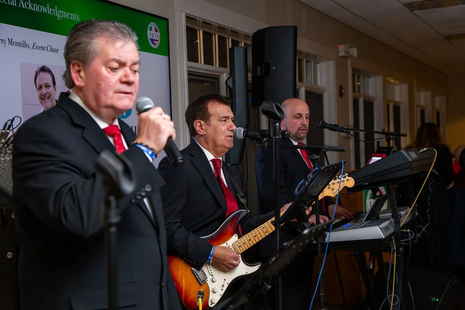 Three men in suits, one holding a microphone, performing with musical instruments, at an indoor event with a green banner in the background.