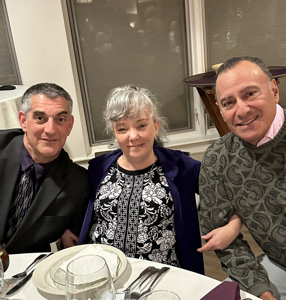 Three people sitting at a dinner table, smiling at the camera. The table is set with plates, glasses, and silverware.