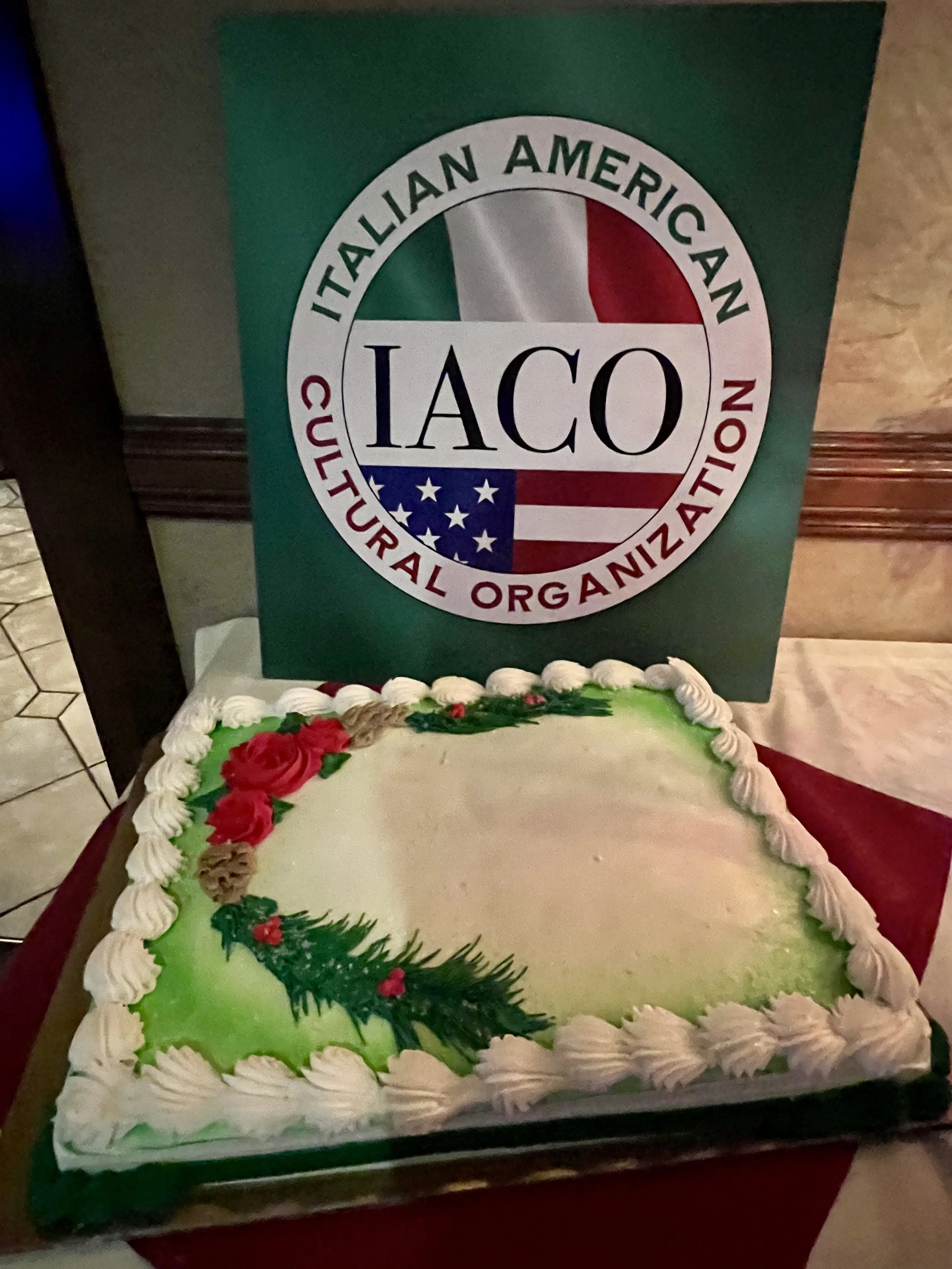 A decorated Christmas-themed cake with green icing, red roses, and a sprig of holly. In the background, there is a sign for the Italian American Cultural Organization (IACO).