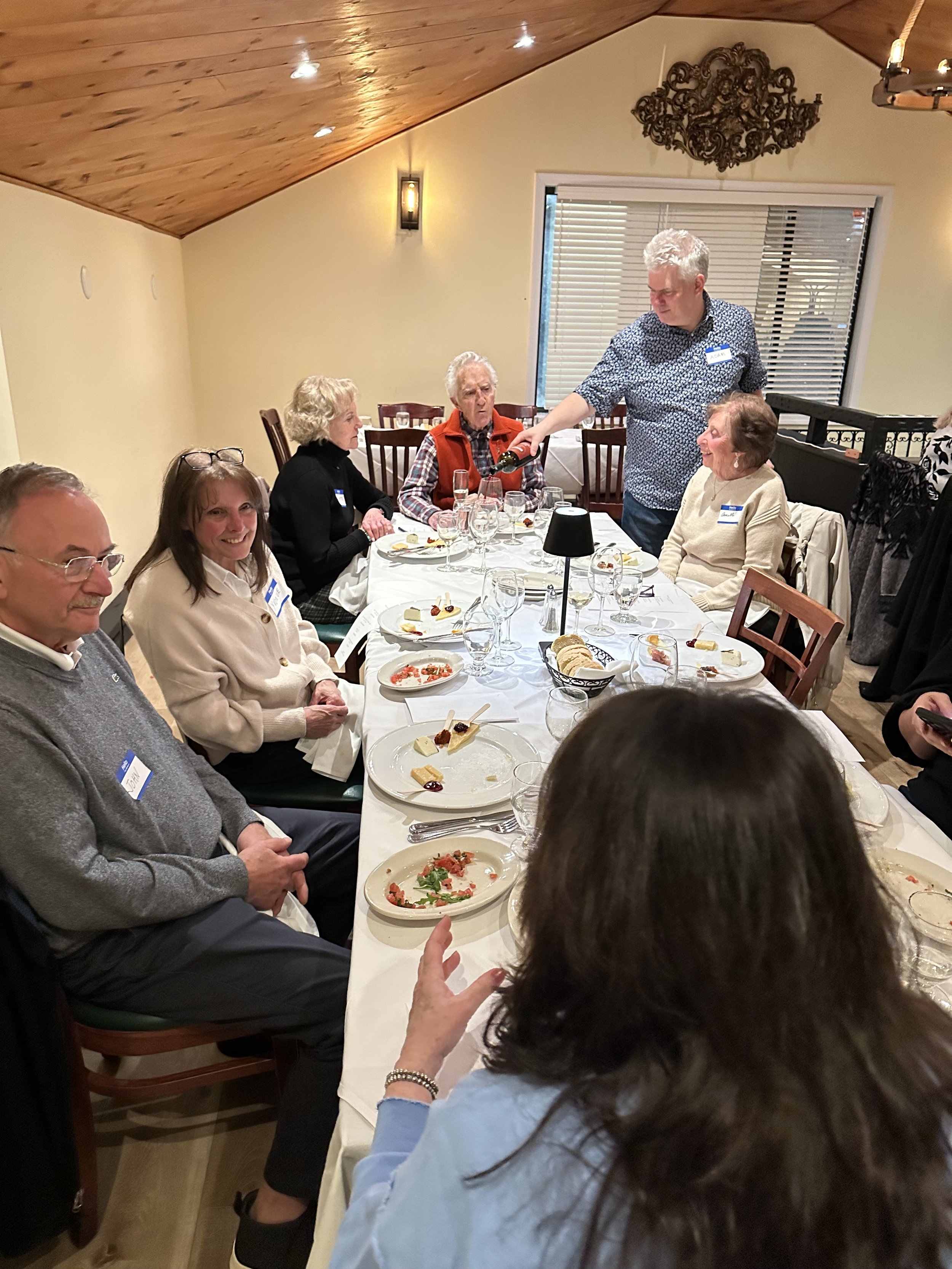 Group of people sitting around a dining table with wine glasses and plates, in a cozy restaurant setting.