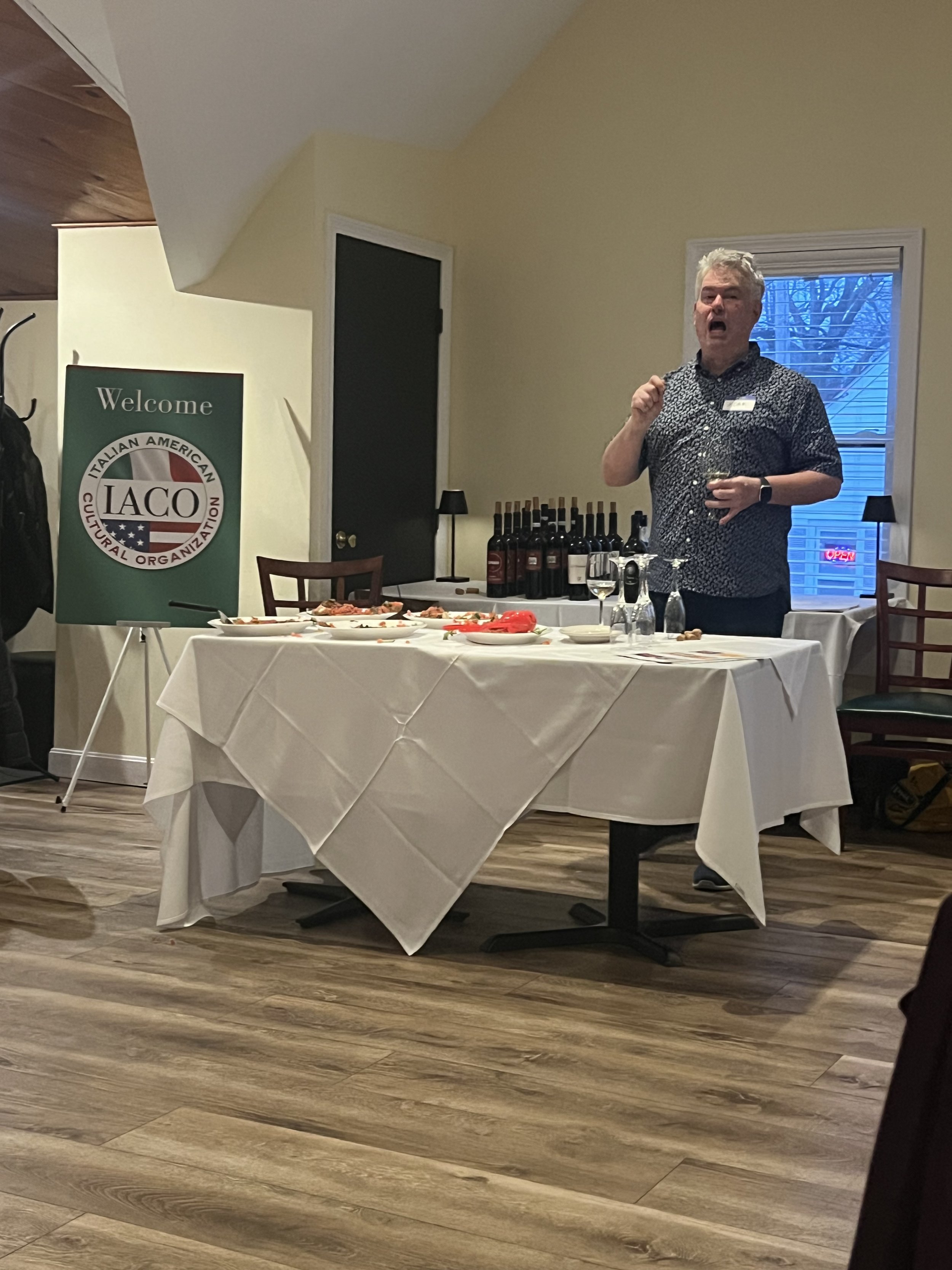 A person speaking in a room with tables set for an event, featuring bottles of wine and a sign that reads 'Welcome Italian American Cultural Organization'. The room has wooden floors and a window in the background.