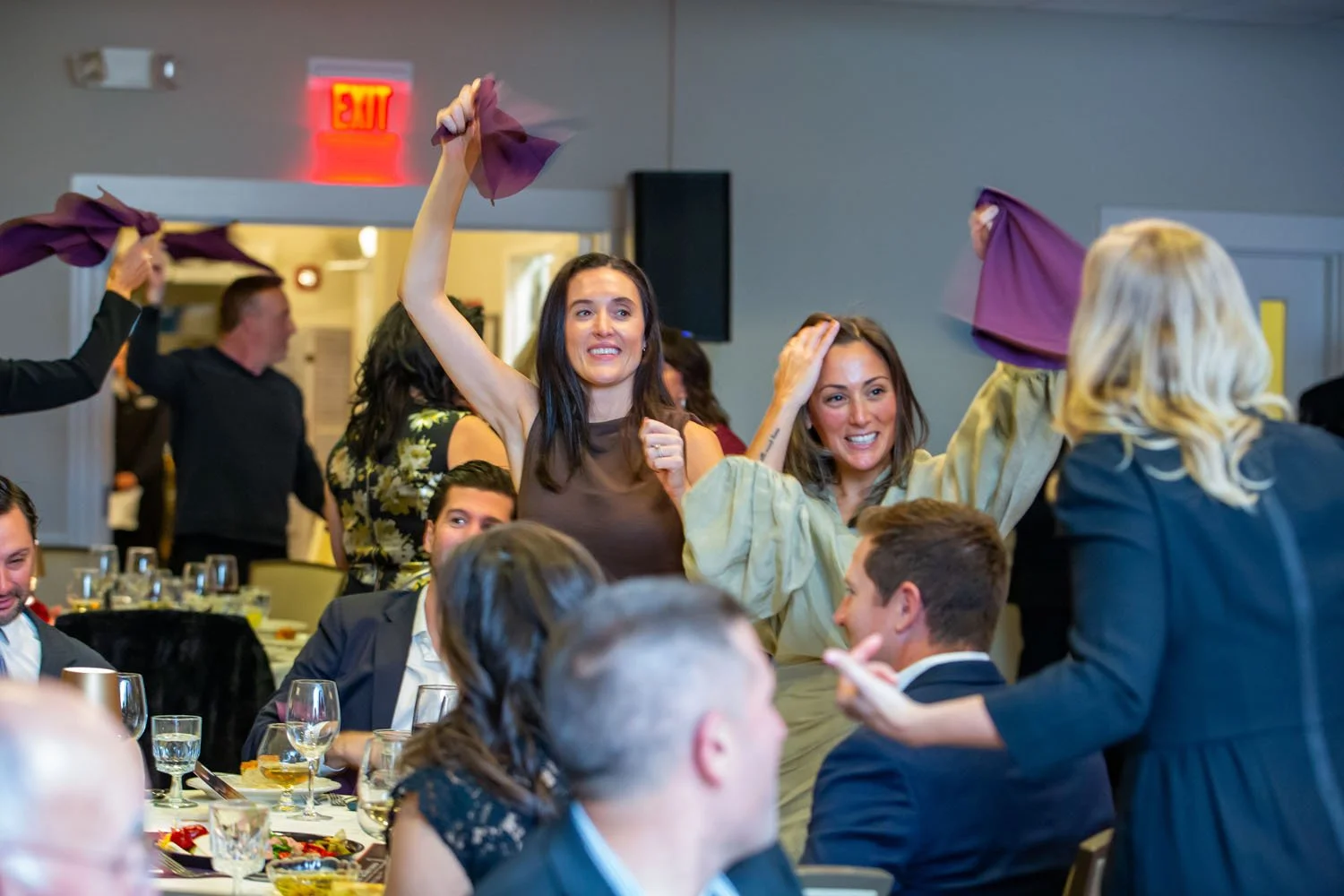 People celebrating at a banquet, with women in Middle Eastern attire dancing and throwing purple cloths, surrounded by seated guests, tables with glasses and plates, in a banquet hall.