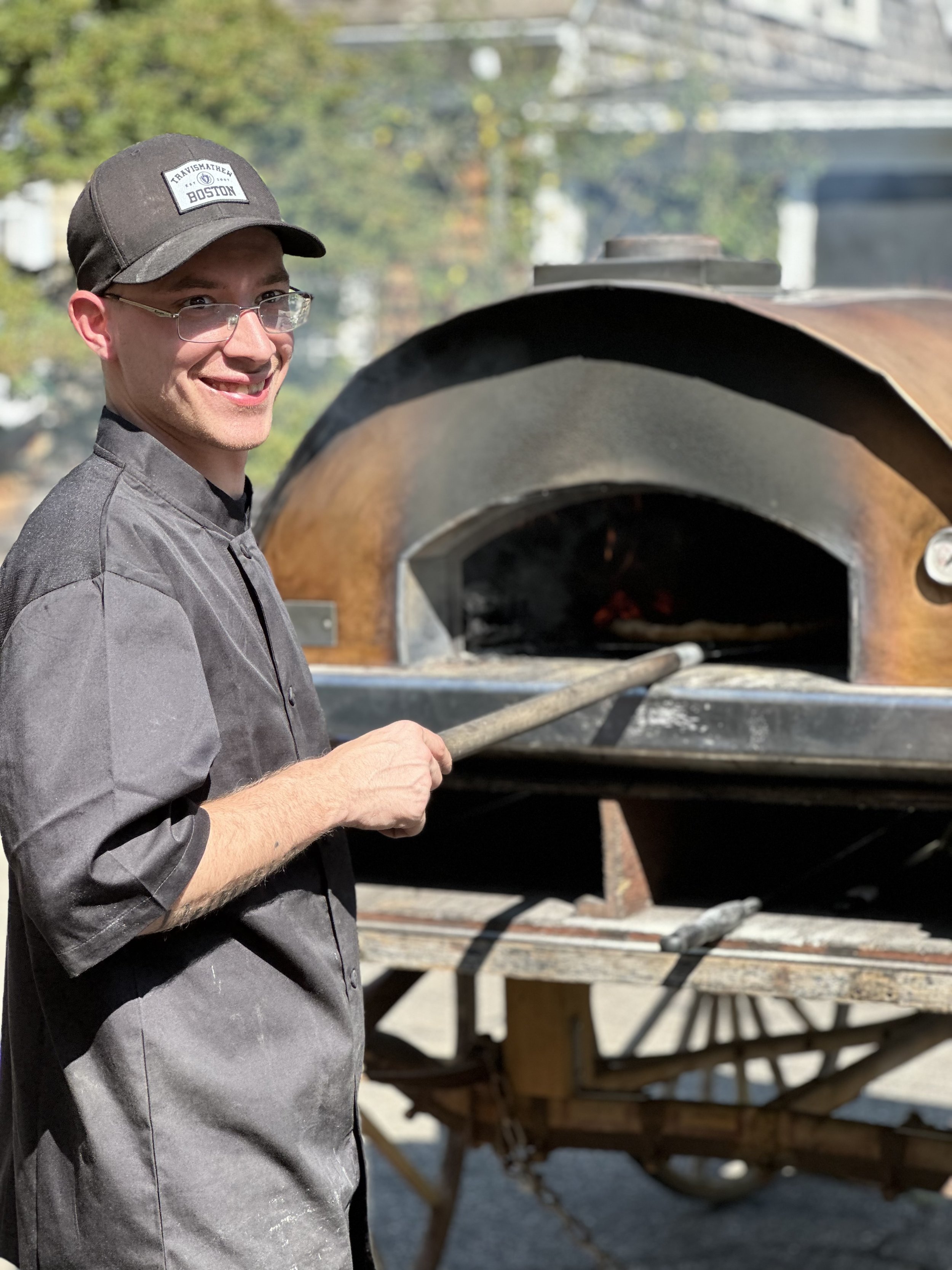 Person in black chef attire and cap smiling next to a wood-fired oven, holding a long-handled tool outdoors.