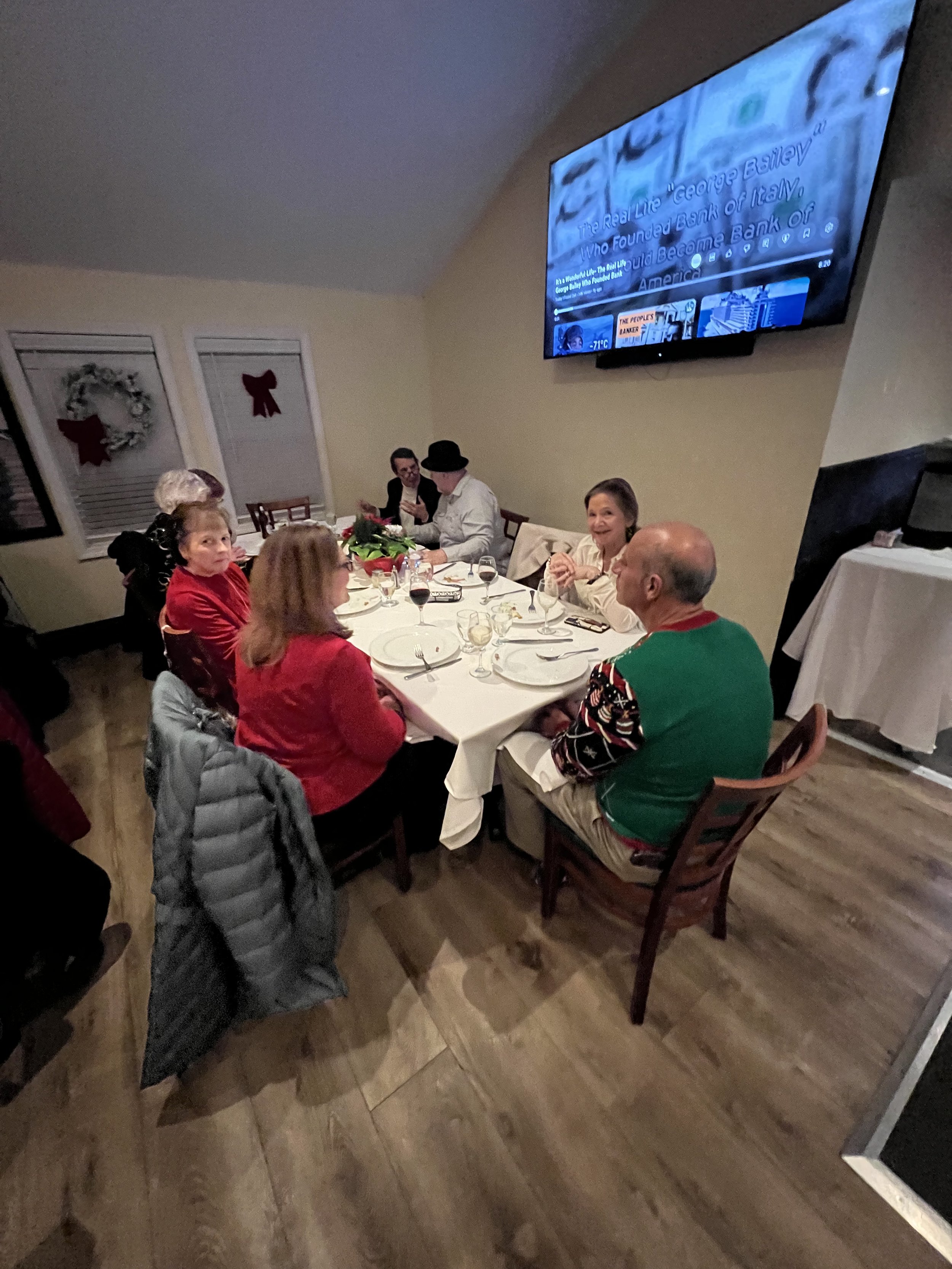 People dining at a restaurant table decorated for a holiday, watching a TV displaying a finance-related program.