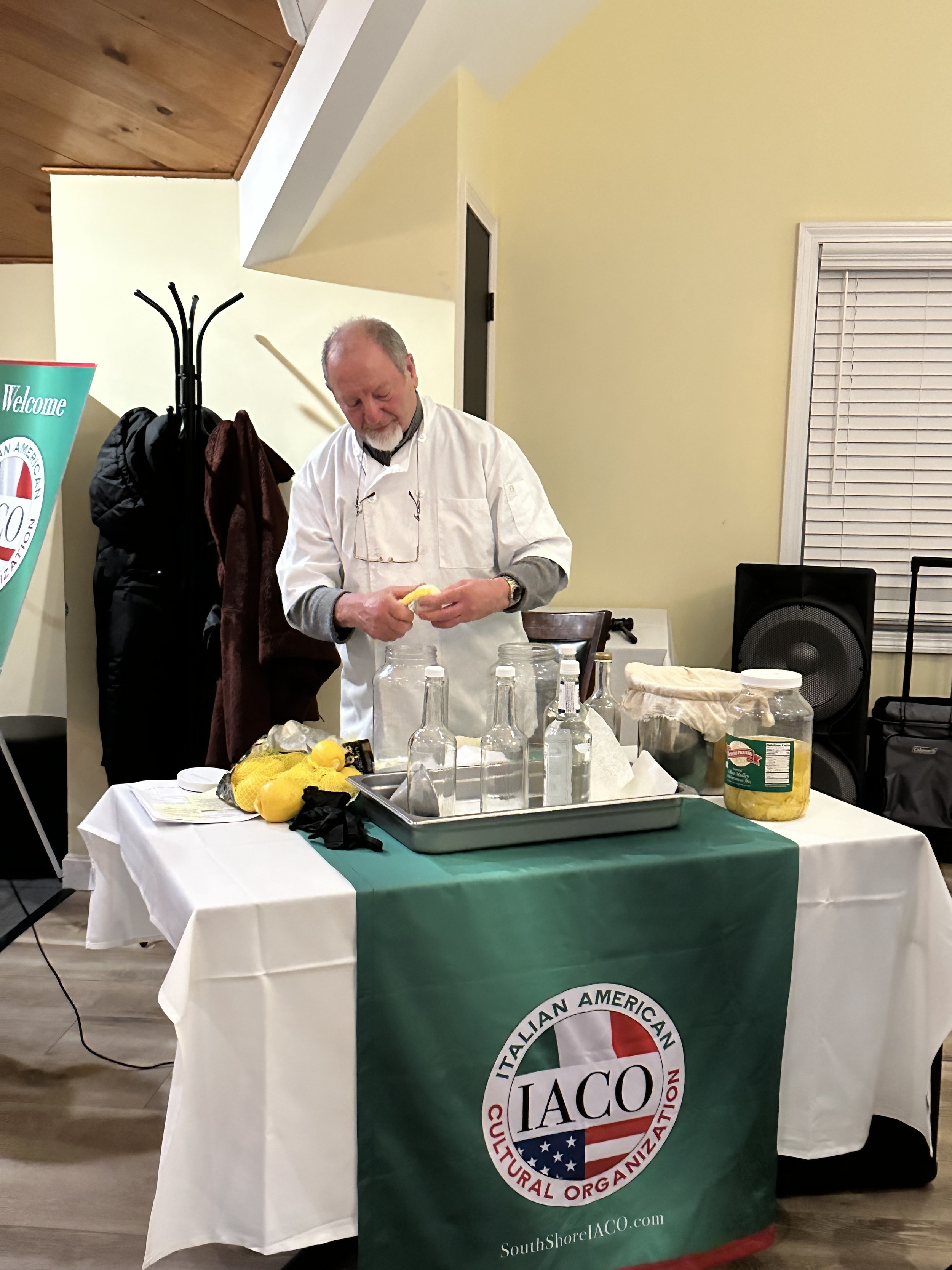 A man peels a lemon at a table with bottles and lemons, draped with a cloth bearing the Italian American Cultural Organization logo.