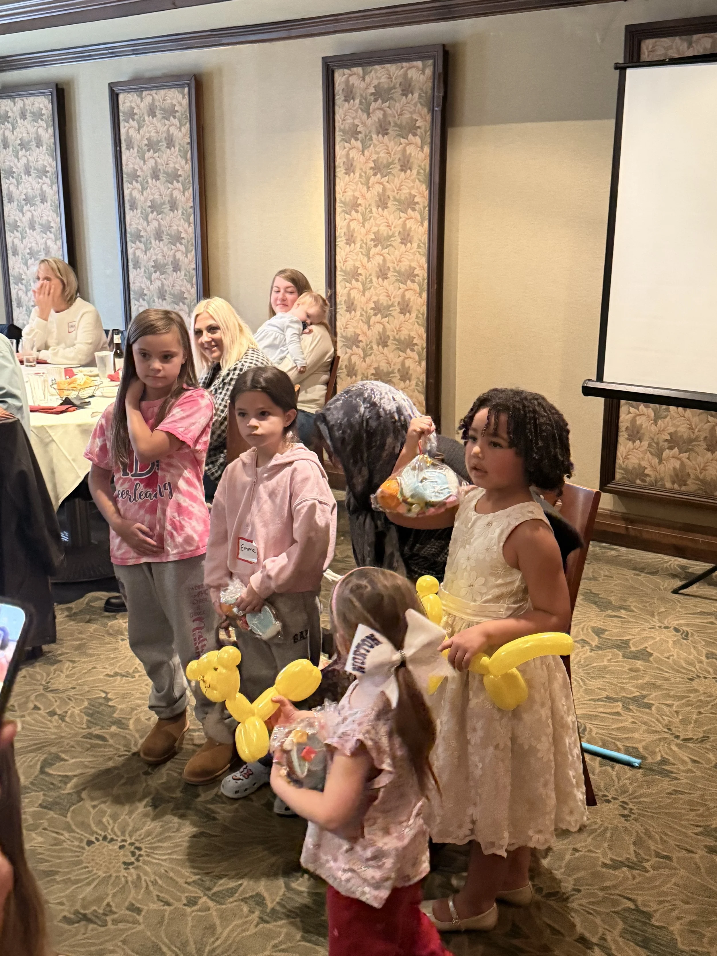 Children gathered around at a birthday party, holding yellow balloon animals, with adults seated at tables in the background.