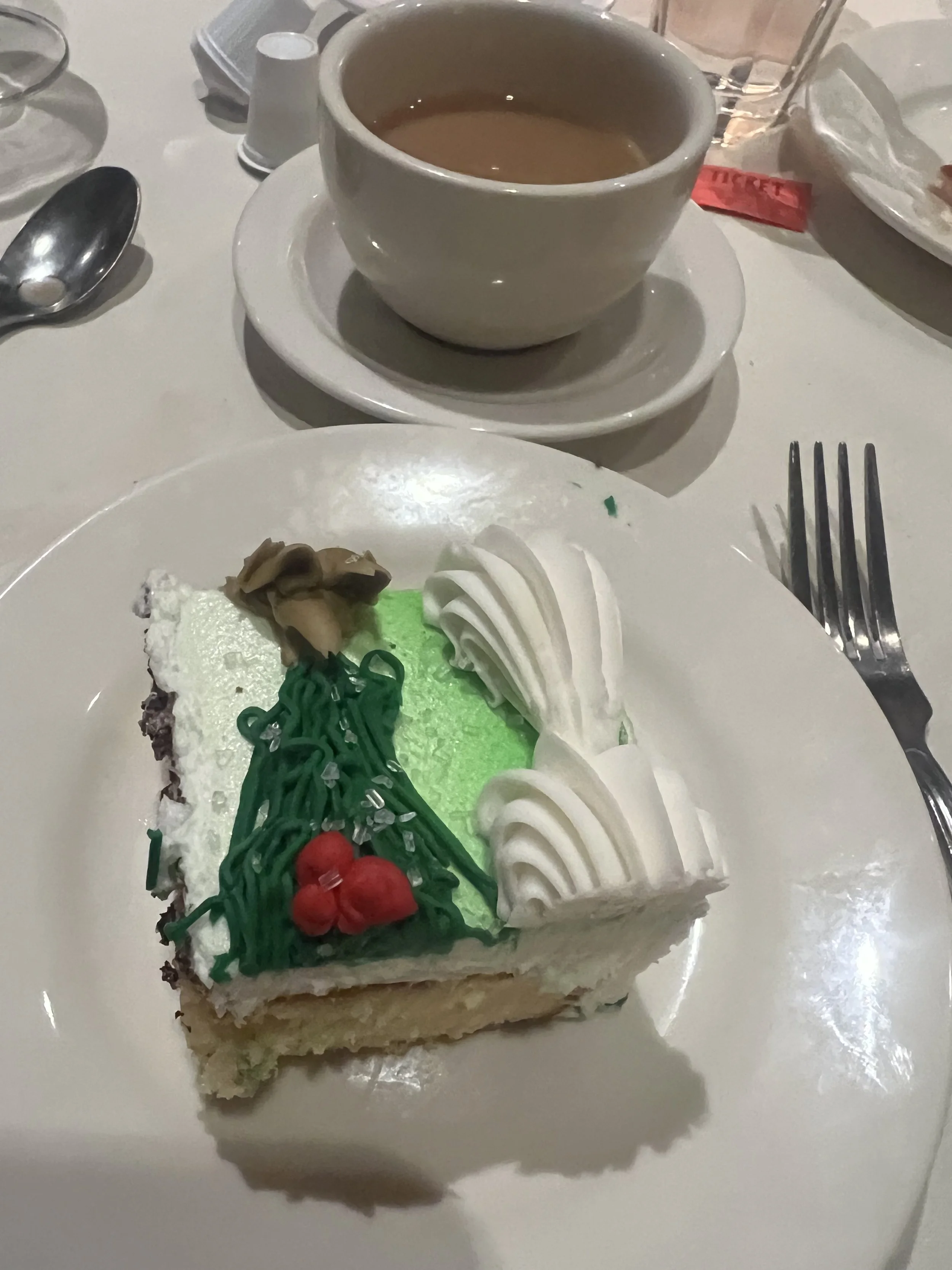 A slice of Christmas-themed cake with white frosting, green and red decorations, and a small cluster of red berries, placed on a white plate. A cup of coffee with milk or cream is set behind the cake, on a saucer. Silverware and glasses are visible a