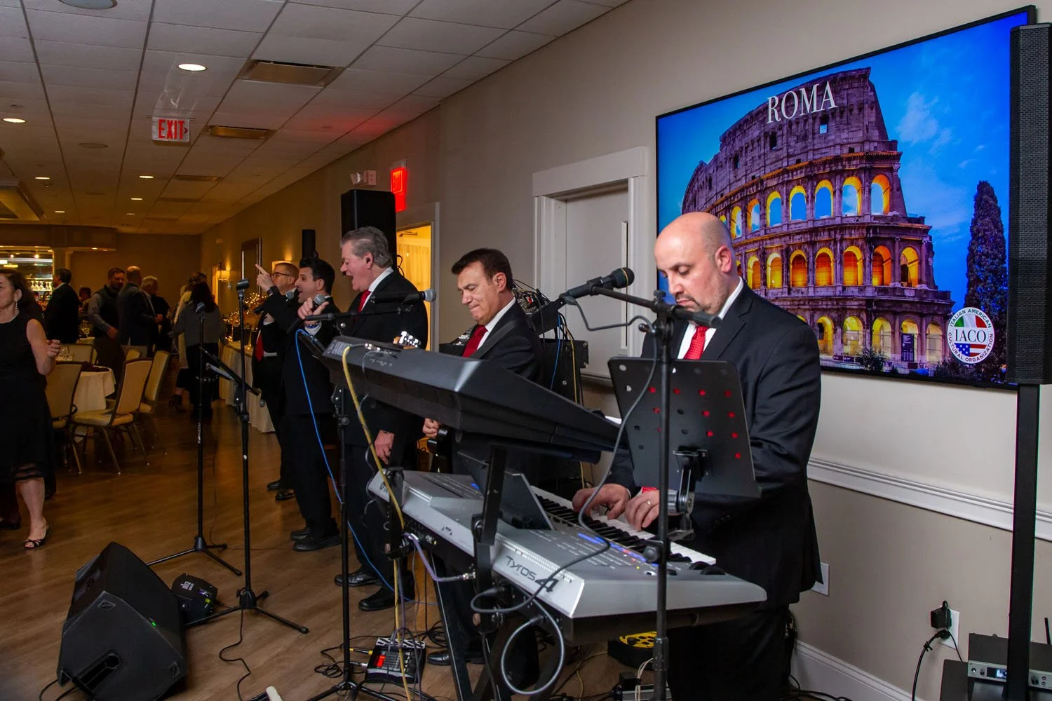 Band playing music at an event, with four musicians in tuxedos, one on keyboard, in front of a large screen showing the Roman Colosseum and a logo.