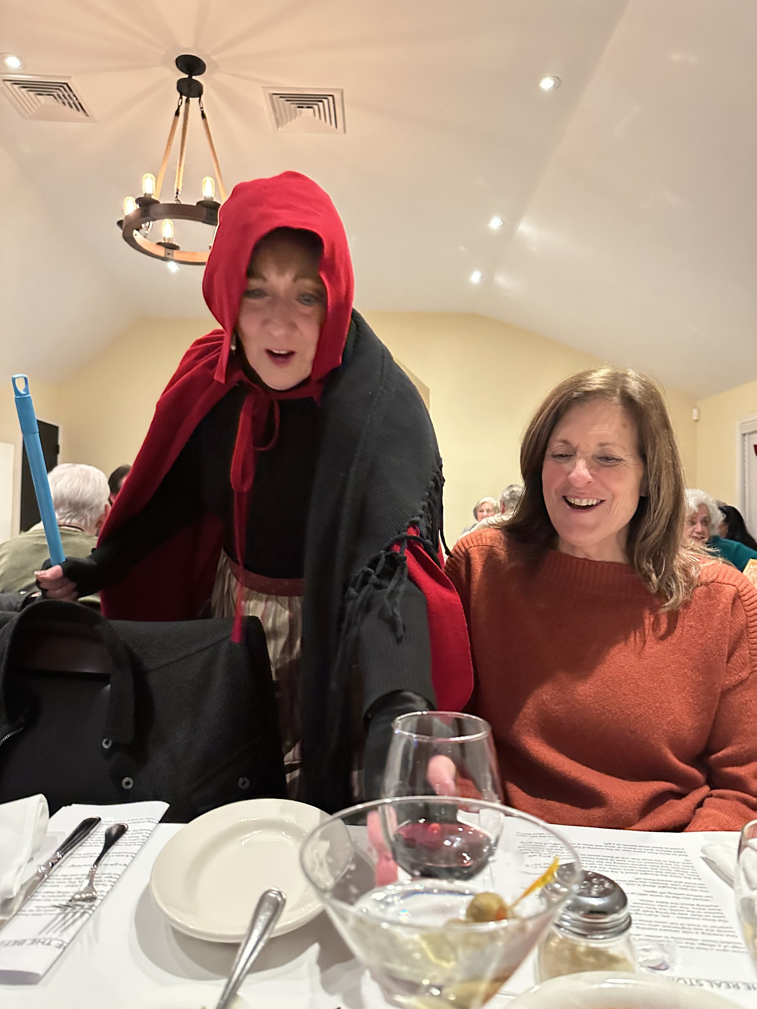 Person in a red hood and black cloak interacting with a seated woman at a dining table, featuring plates and glasses, in a warmly lit room.