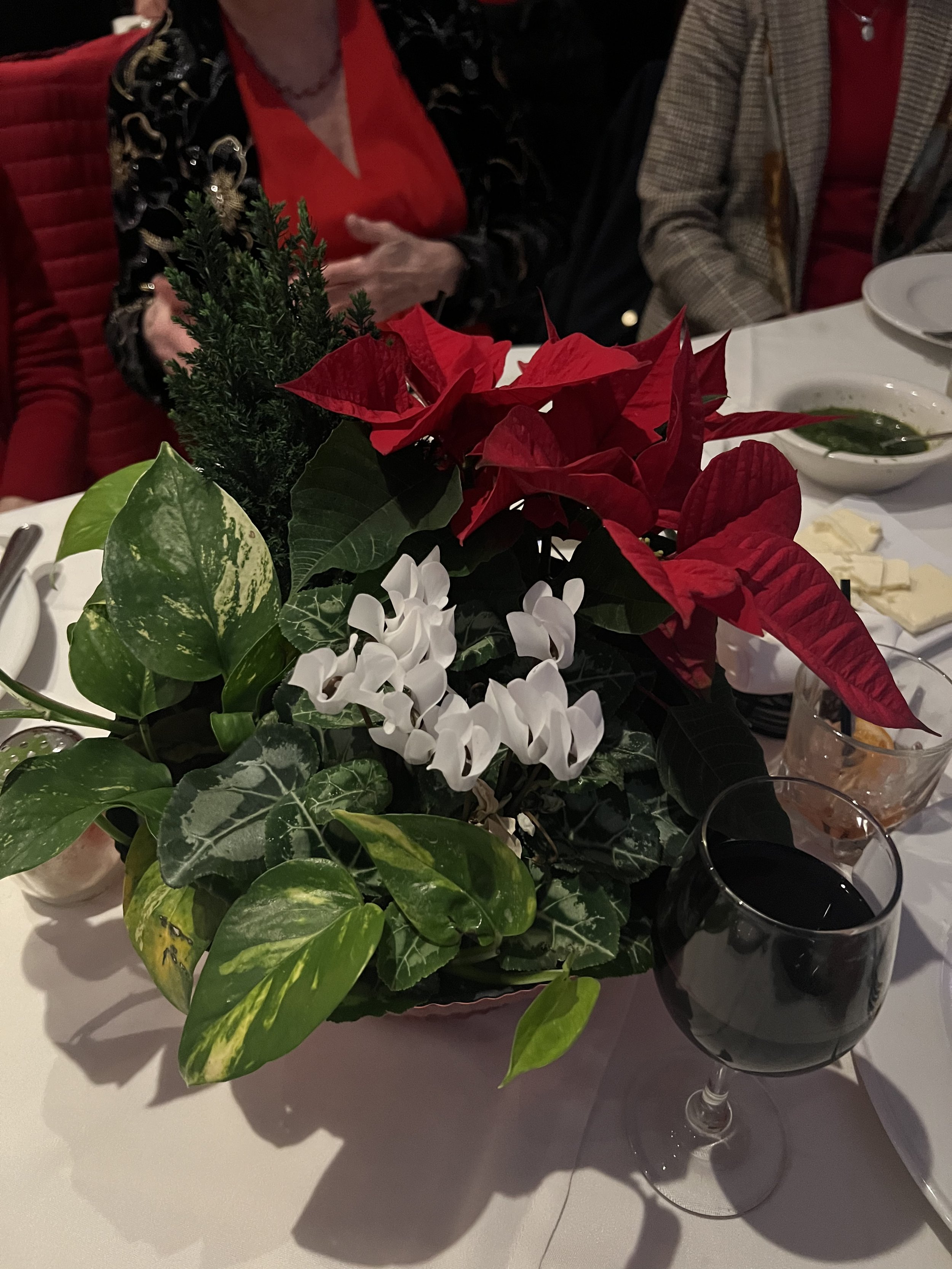 Table centerpiece with red poinsettias, white flowers, green leaves, a glass of red wine, and people in festive attire.