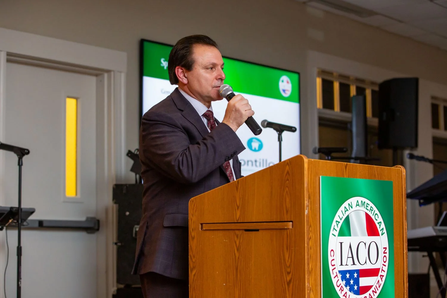 A man in a suit speaking into a microphone at a podium with a green and white logo that reads Italian American Cultural Organization.
