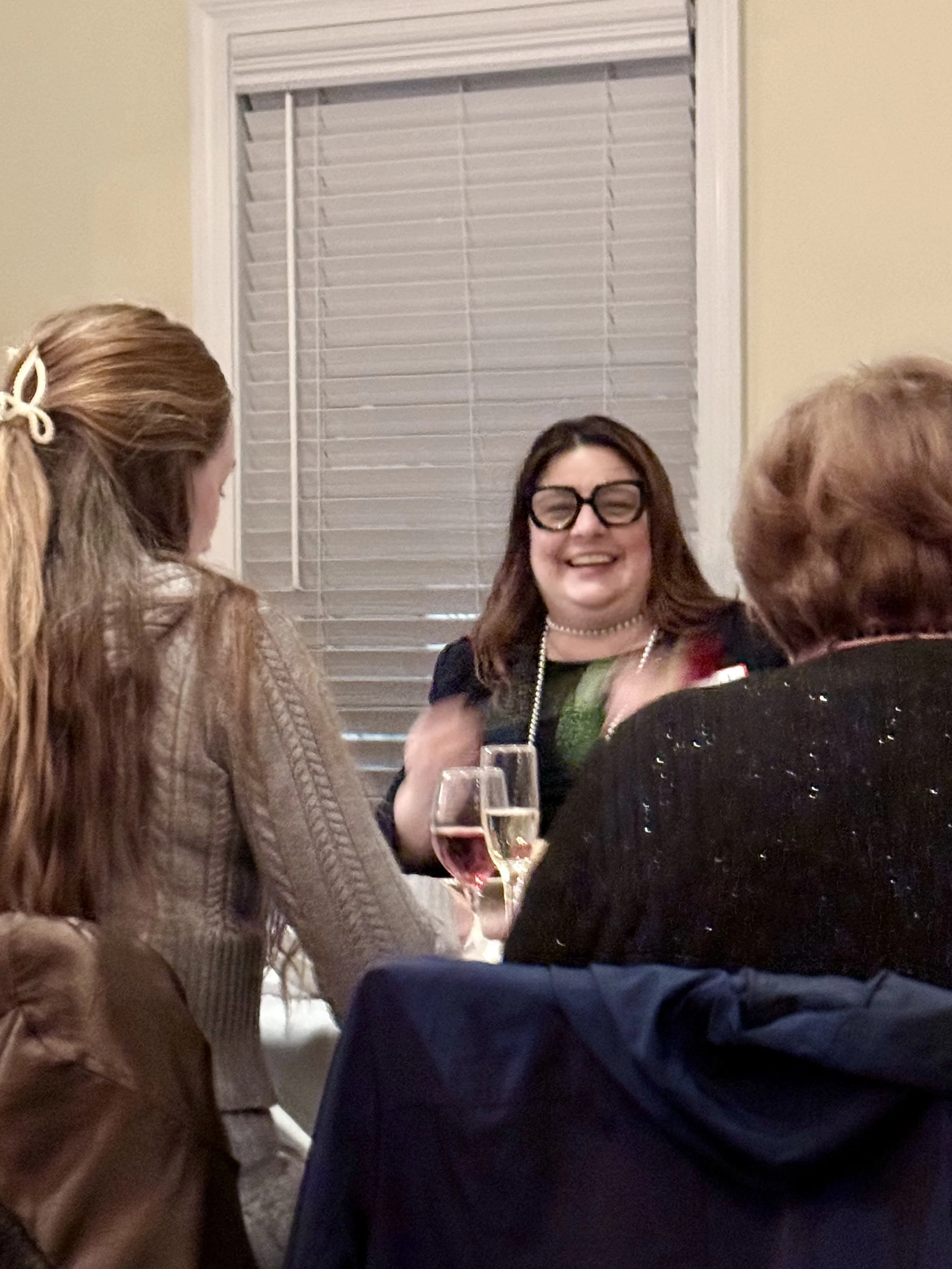 Three women sitting at a table having a conversation, with two glasses of champagne in front of them, inside a room with closed blinds.