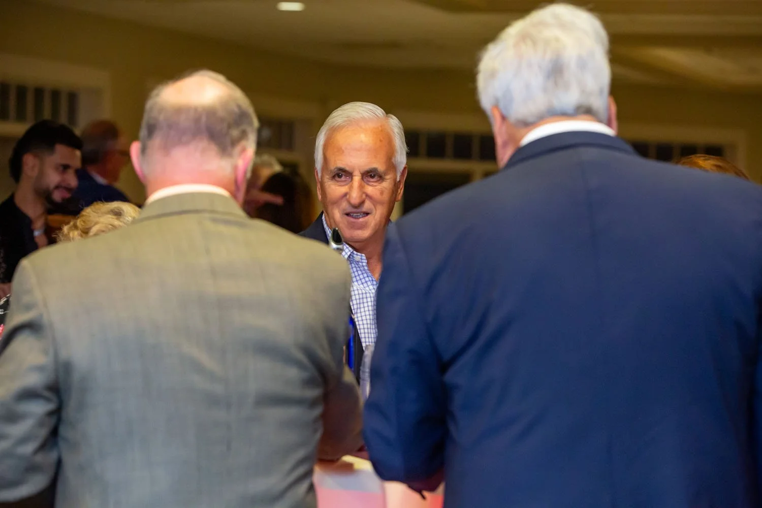 Three men engaged in conversation at a social event, with one man smiling in the center, facing two others with their backs to the camera.